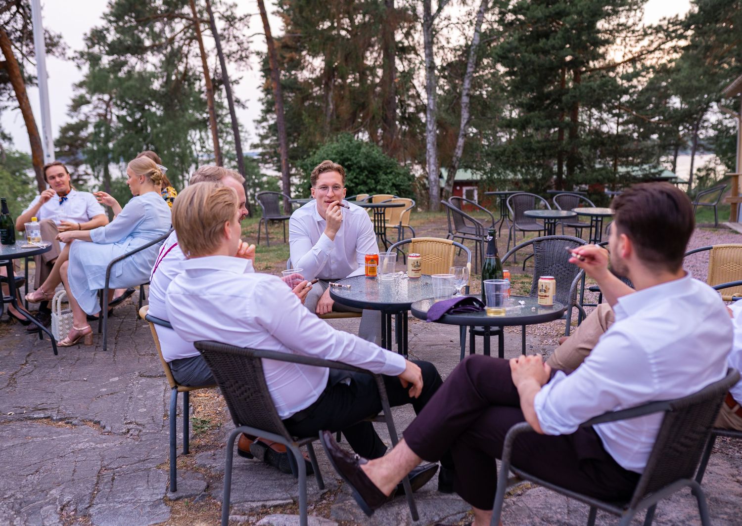 Wedding guests socializing at outdoor patio tables during evening reception surrounded by tall pine trees.