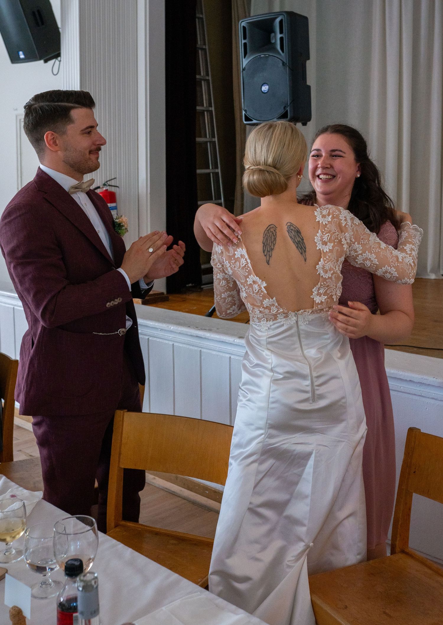 Joyful bridesmaid in pink hugging the bride in a lace wedding gown at the reception.