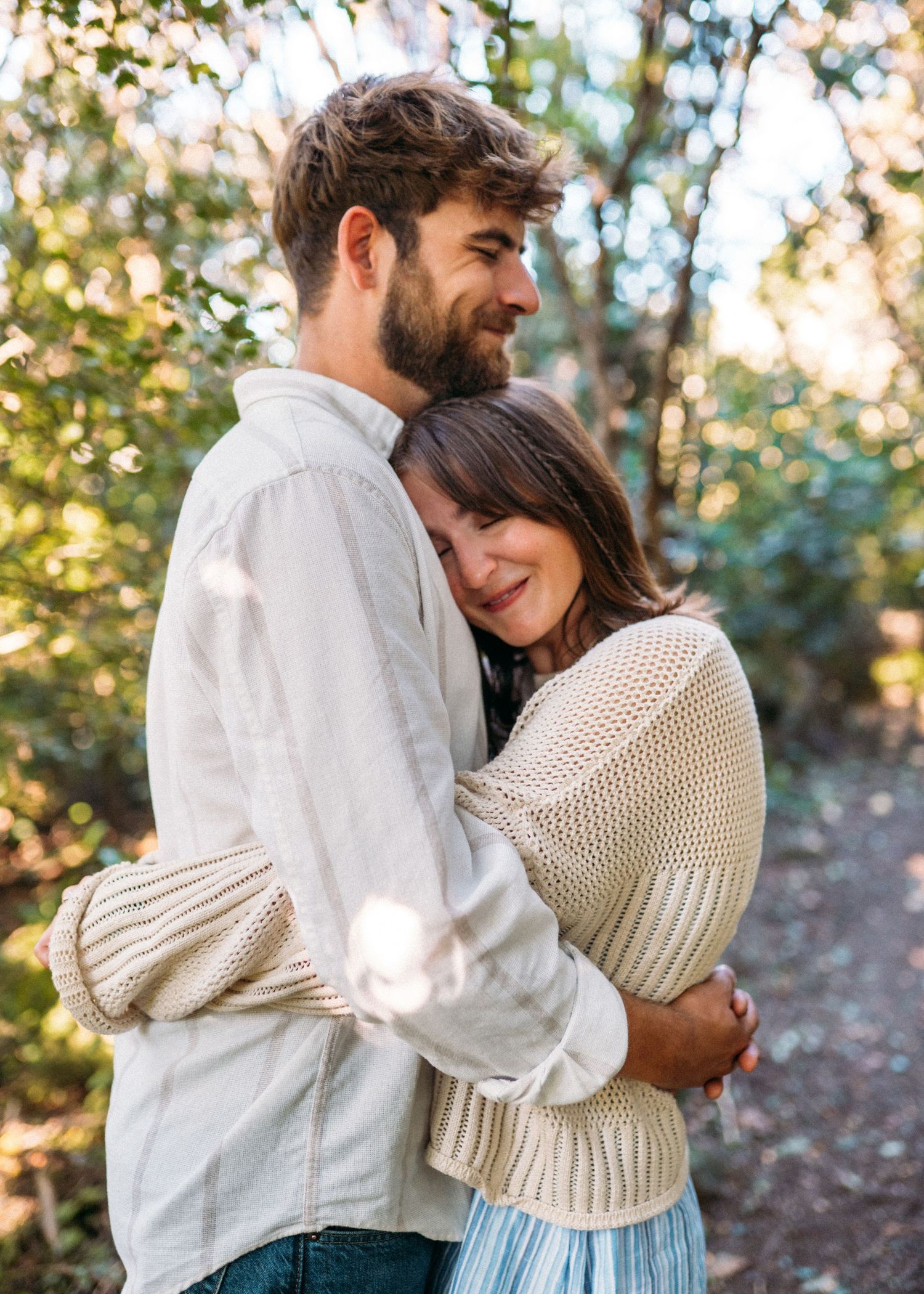 Couple sharing a tender embrace in a sun-dappled forest, woman smiling against mans chest.
