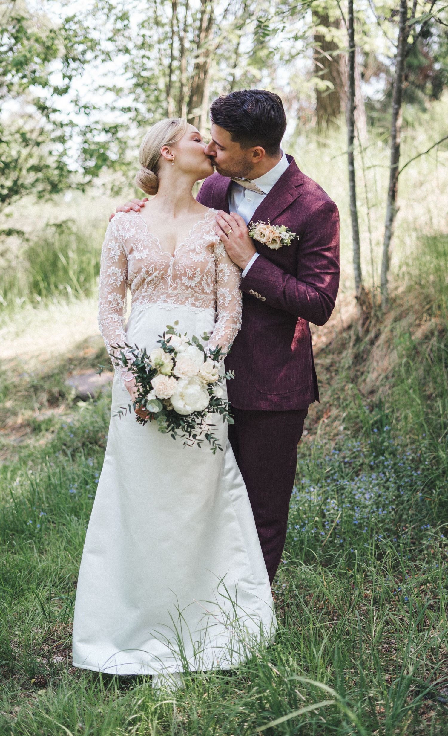 Wedding couple kissing in lush green meadow, bride in lace gown holding white peonies, groom in burgundy suit.