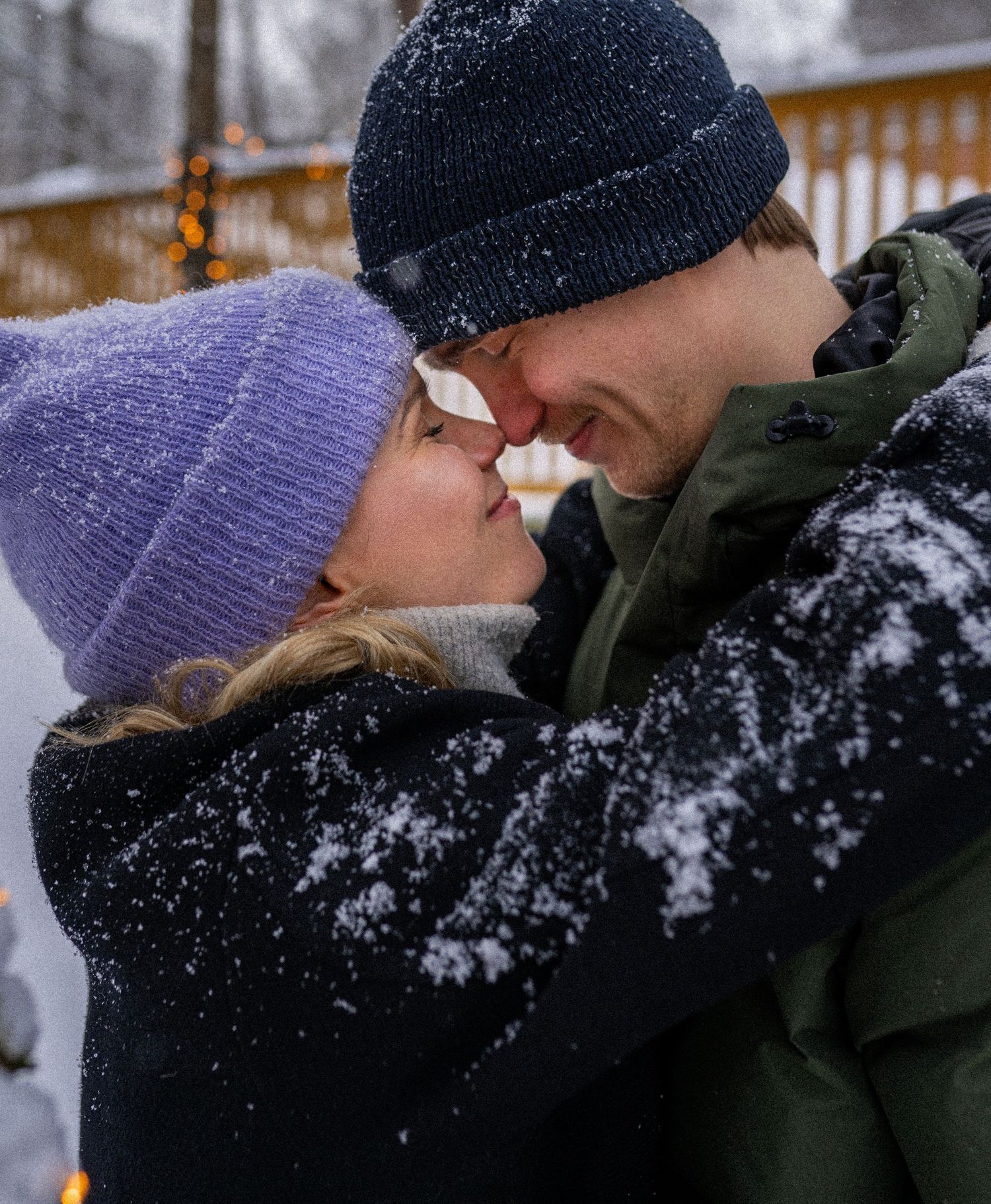 A couple sharing a romantic kiss in the snow, wearing winter hats and cozy jackets outdoors.