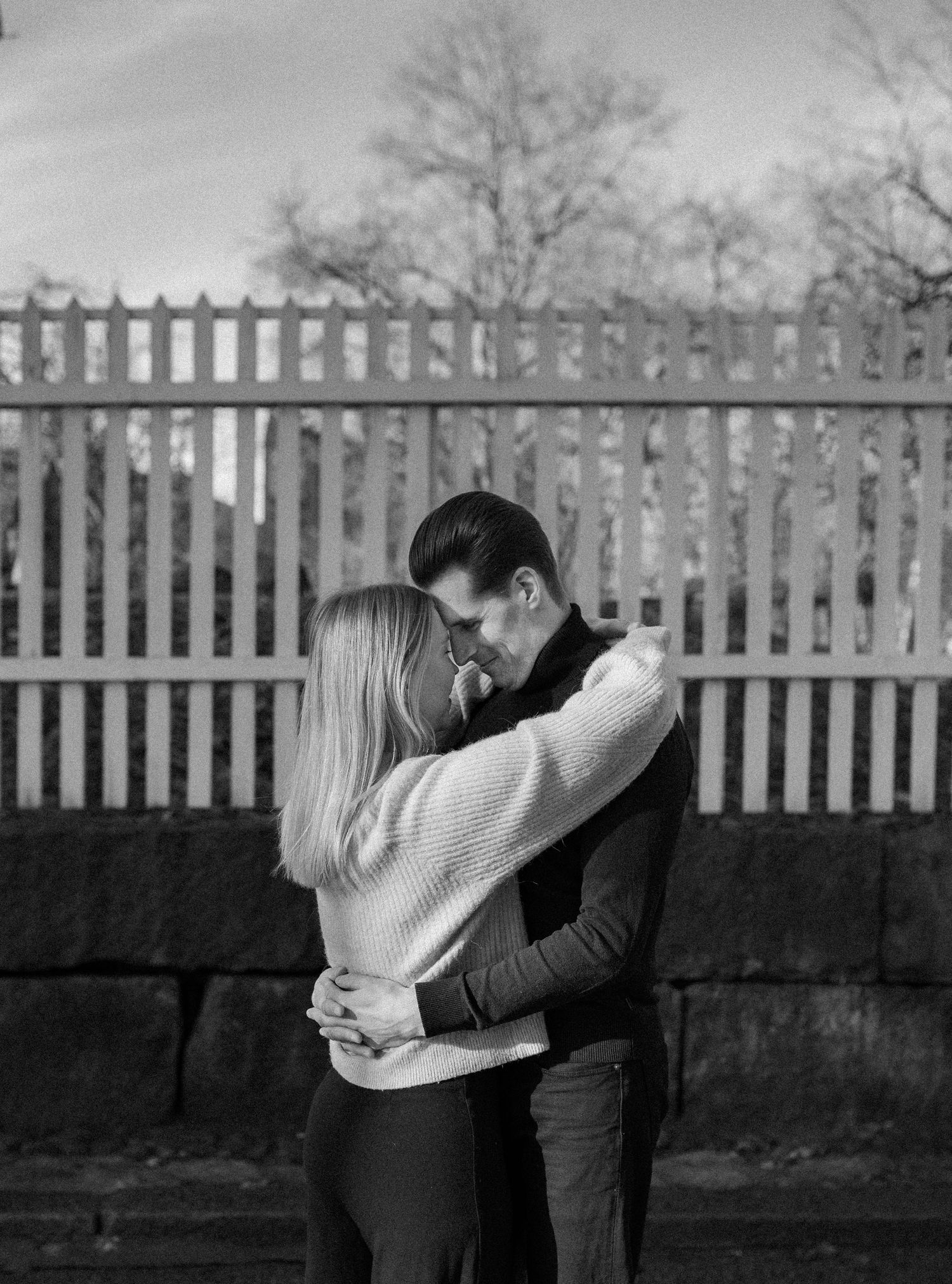 Black and white photo of a couple embracing tenderly outdoors near a stone wall and wooden fence.