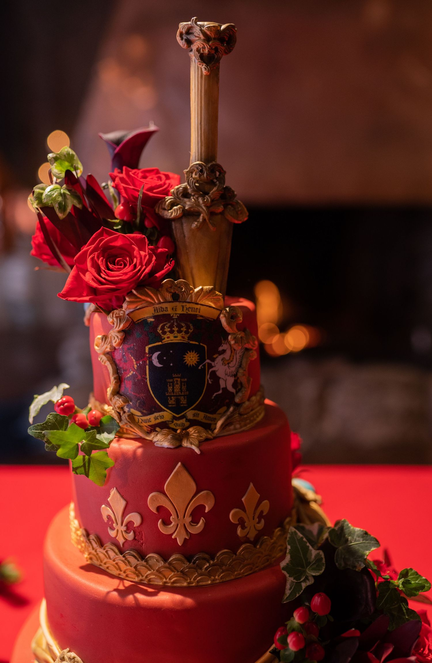 Elegant red royal wedding cake with gold fleur-de-lis, decorative sword, crest, and red roses on a dramatic lit background.