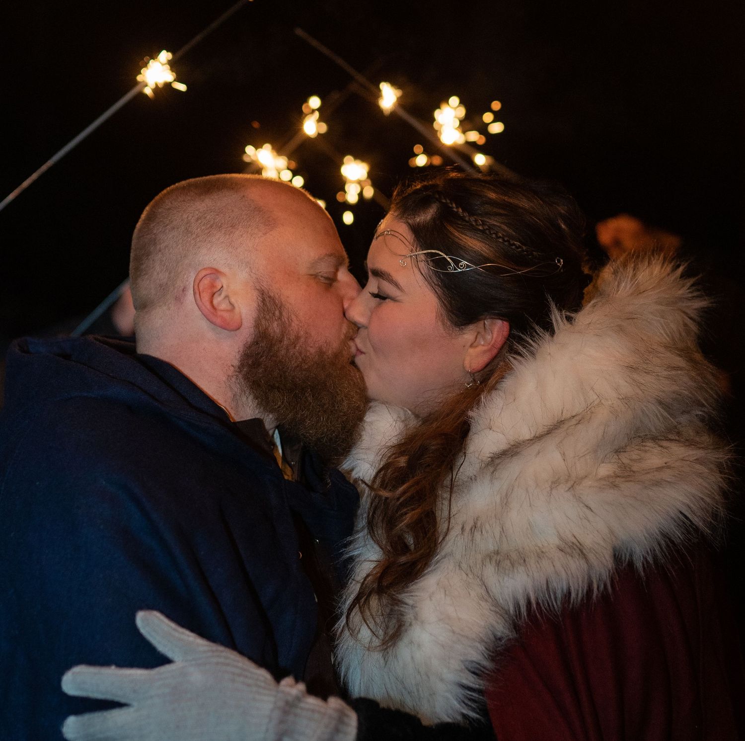 Couple sharing a romantic kiss under sparklers at night, bride in fur wrap and groom in blue jacket.