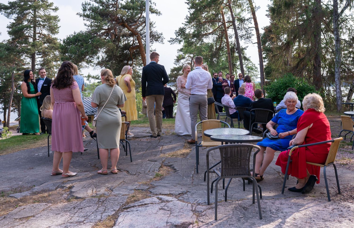 Wedding guests mingling outdoors on a rocky terrace with scenic pine trees and water views.