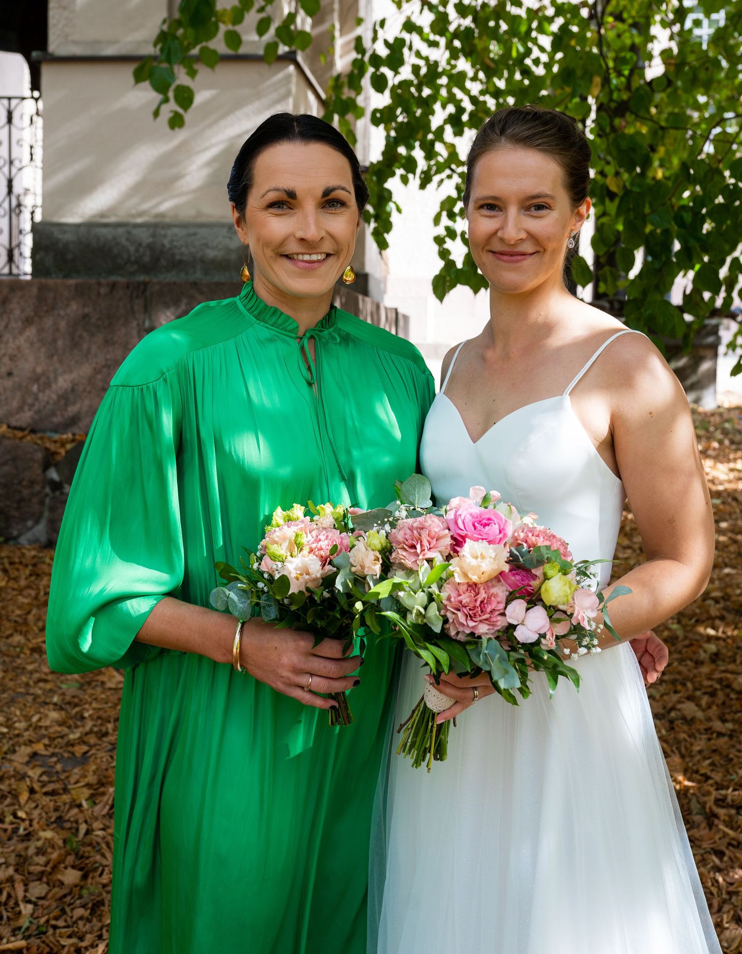 Bride in white gown and woman in green dress posing together outdoors, both holding pink floral bouquets.