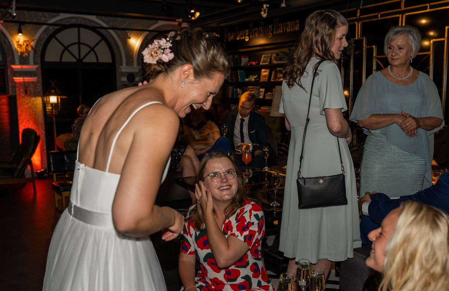 Bride in white dress smiling and chatting with seated guest at wedding reception venue.