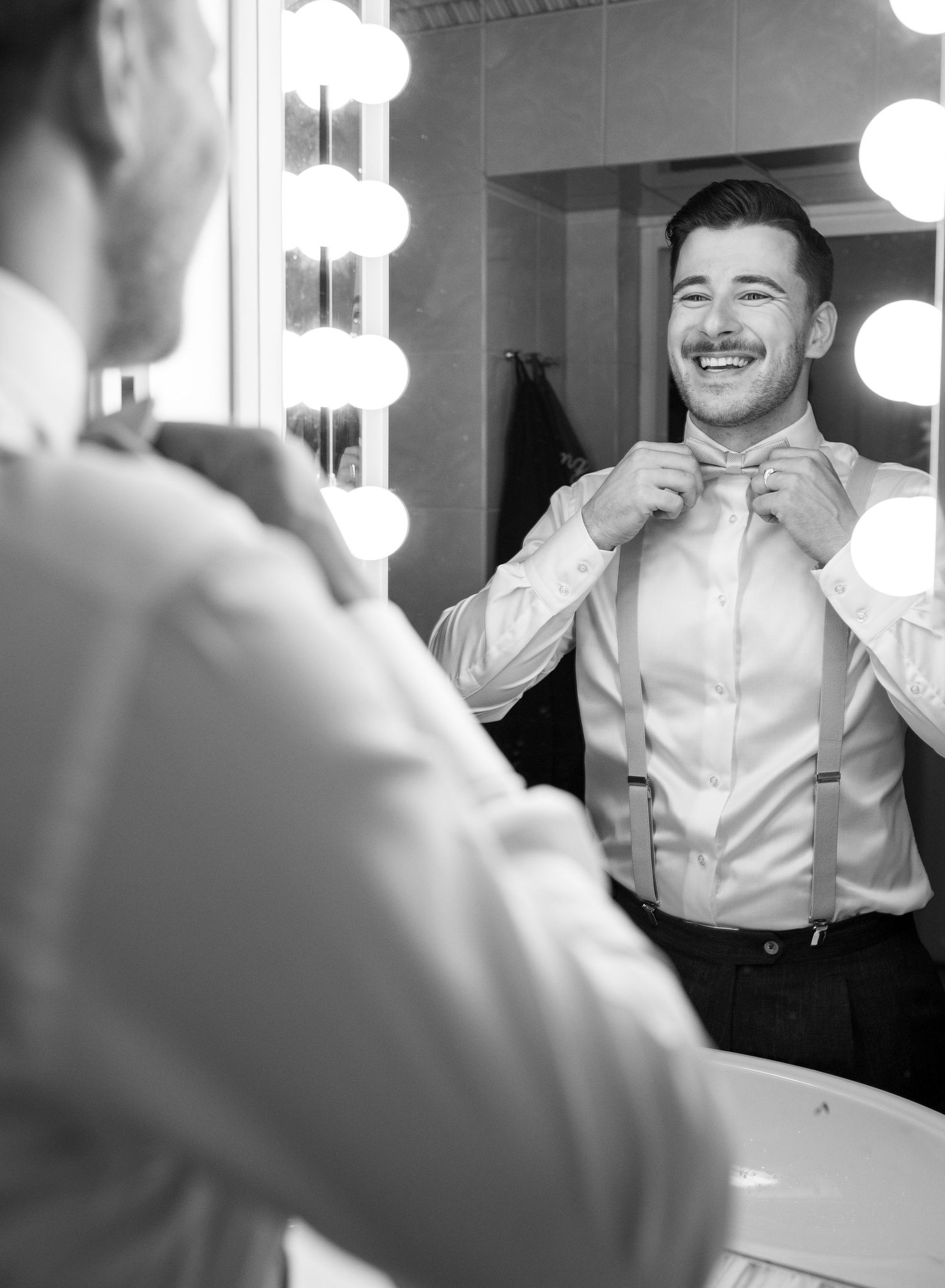 Groom in suspenders smiling at his reflection in a vanity mirror while adjusting his bow tie.