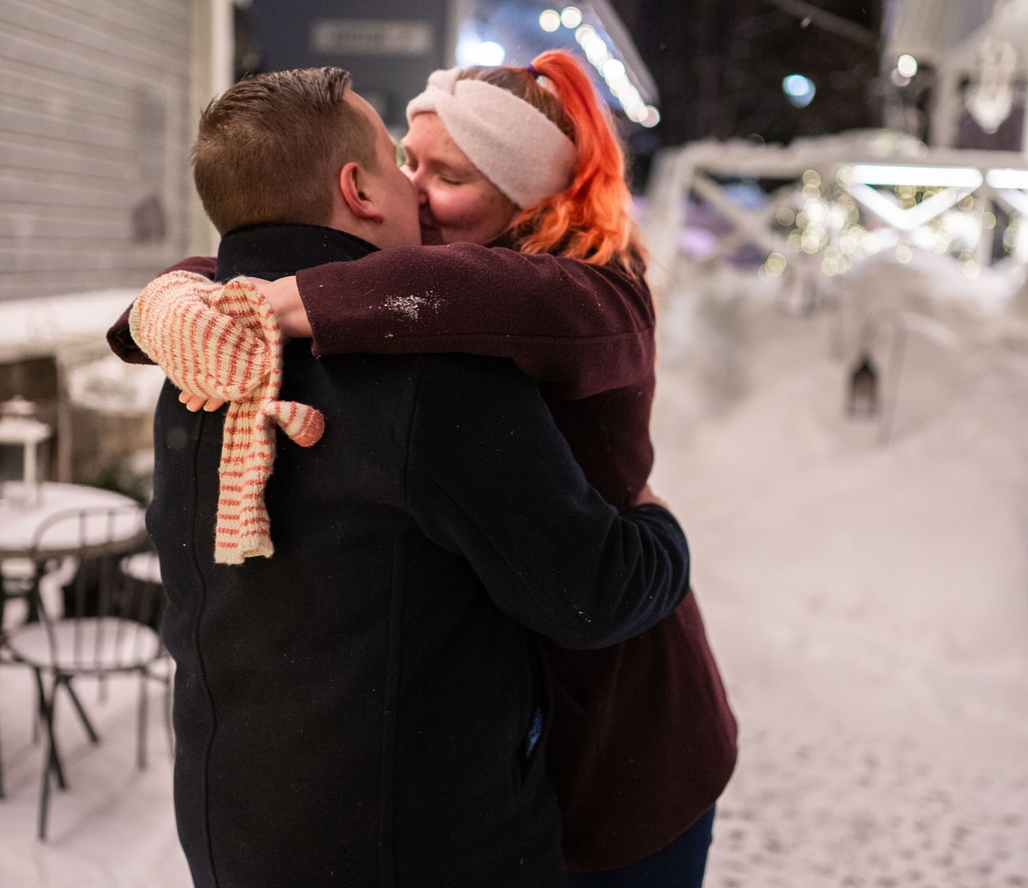Couple sharing a romantic kiss outdoors in a snowy winter setting, woman wearing Santa hat with orange hair.