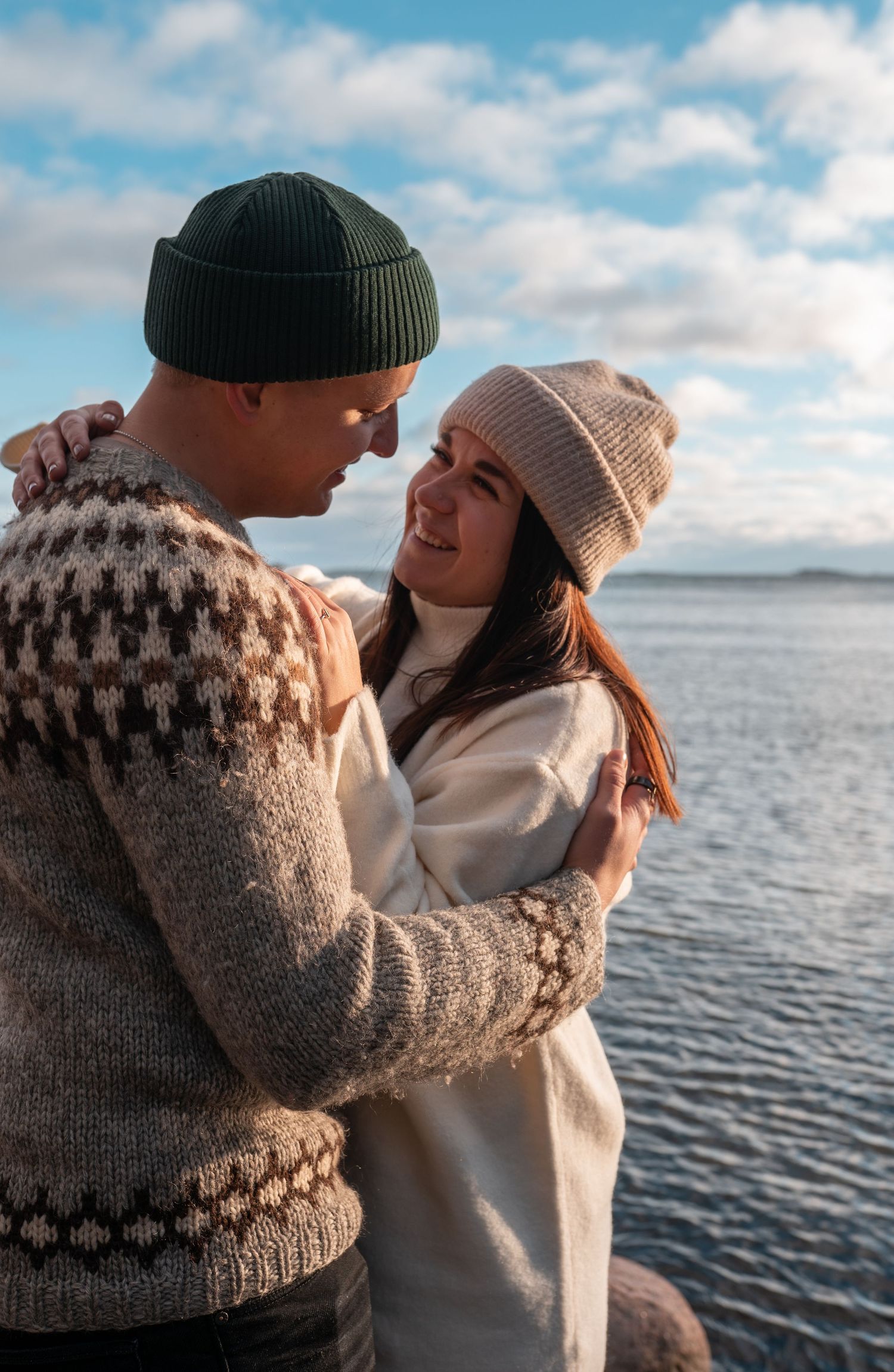 Smiling couple in cozy knit sweaters and beanies embrace by the seaside under a partly cloudy sky.