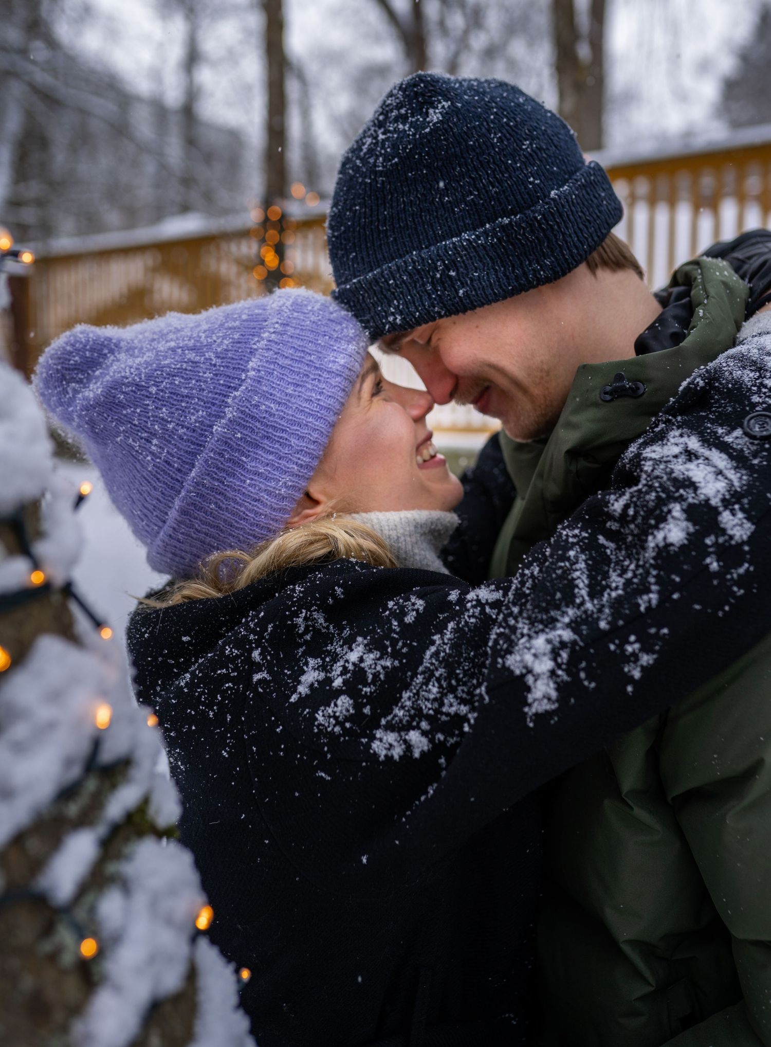 Couple sharing a kiss in snowy winter setting near Christmas lights, wearing colorful beanies and winter coats.