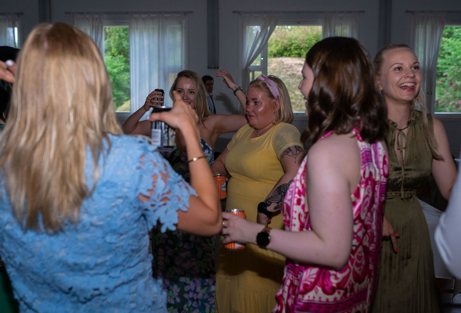 Wedding guests laugh and dance together near large windows during a lively outdoor-lit reception party.