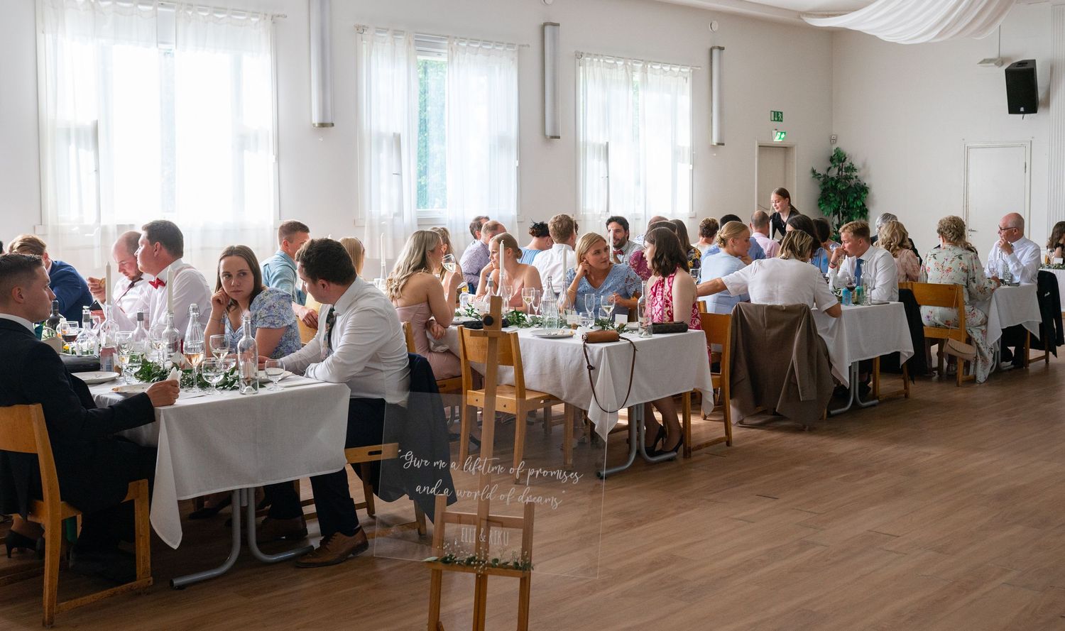 Elegant wedding reception dinner with guests seated at white-clothed tables in a bright hall.