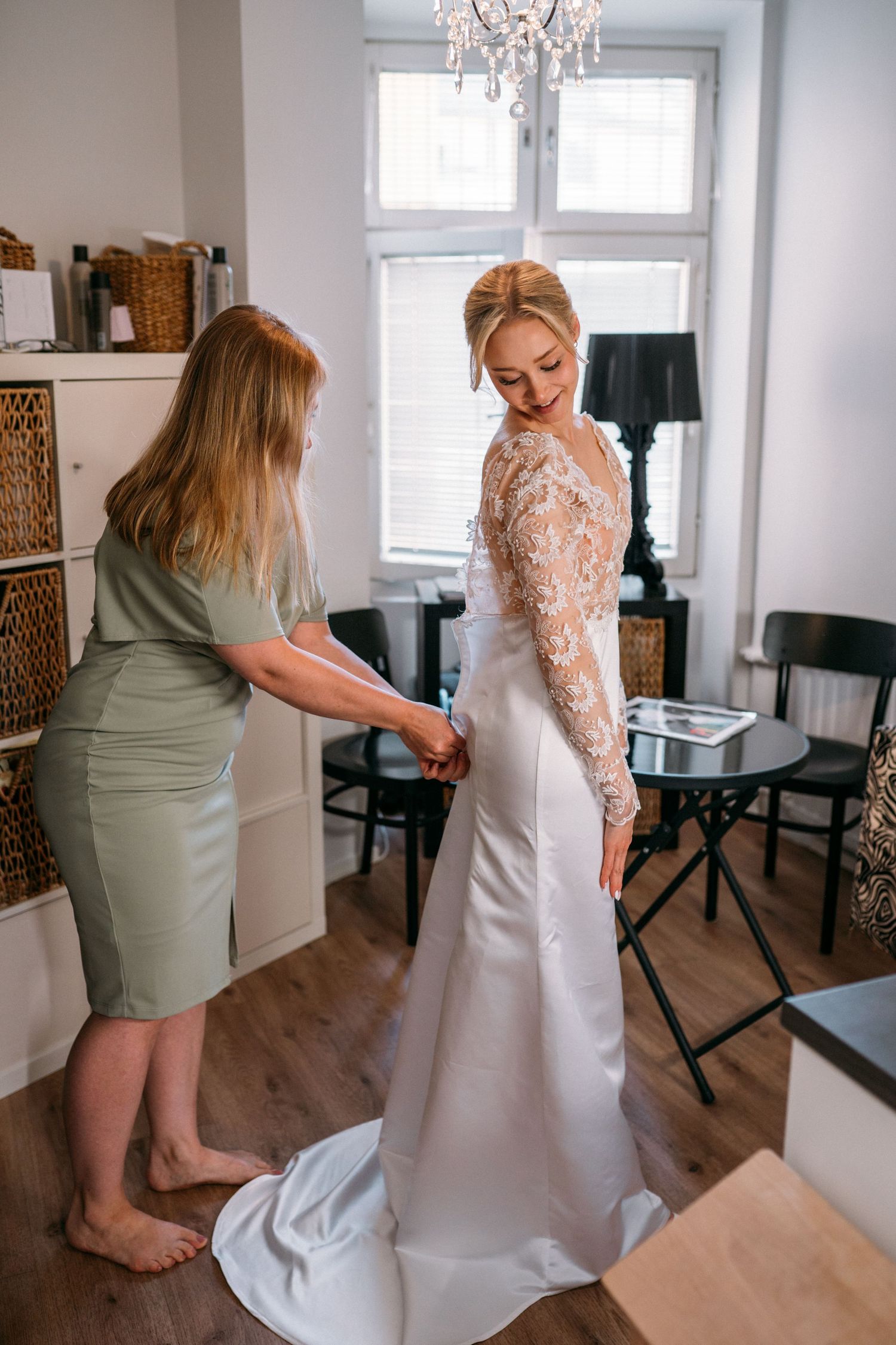 Woman in green dress helping blonde bride button up her elegant lace wedding gown indoors.