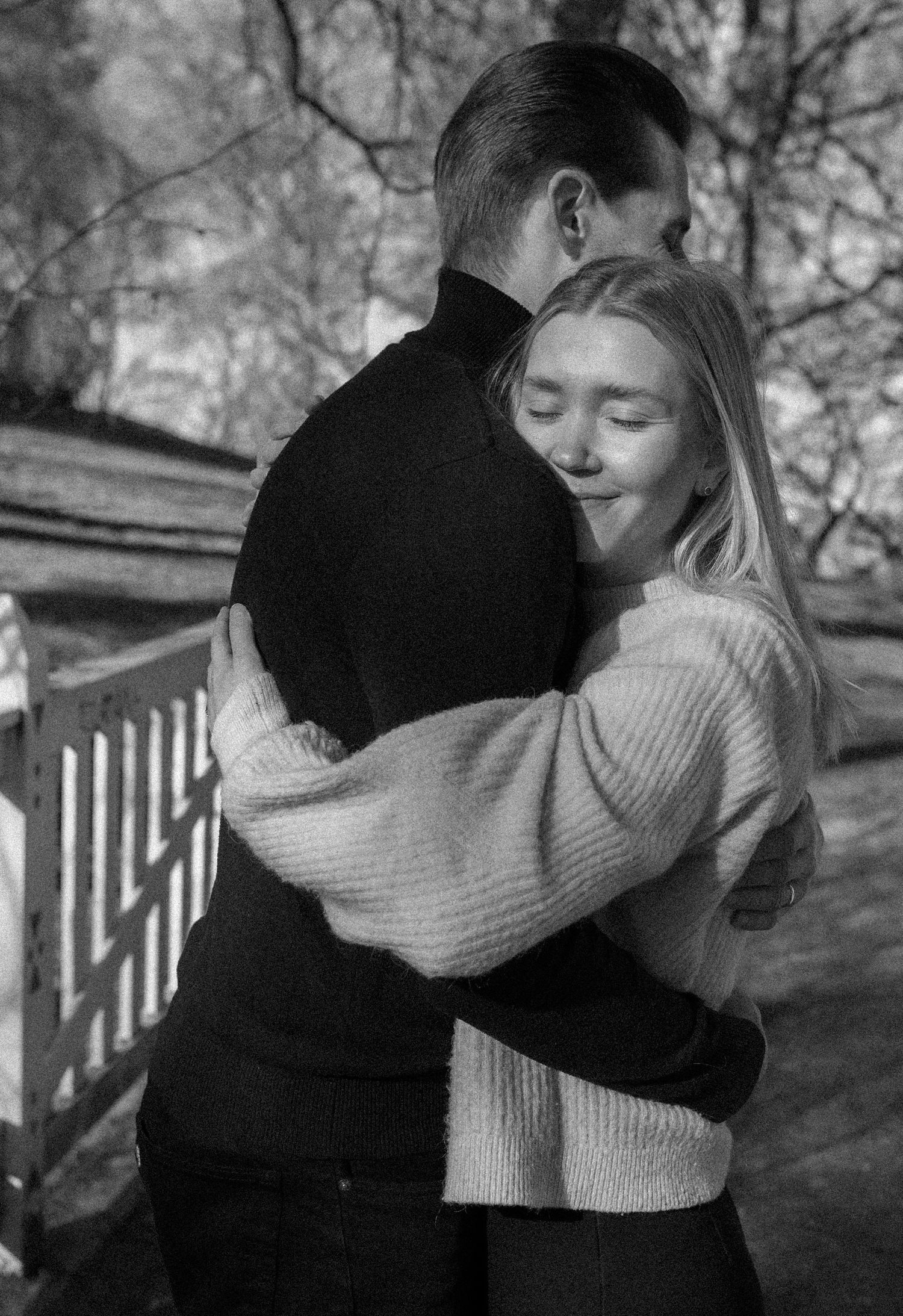 Black and white photo of a couple embracing outdoors, woman in knit sweater smiling with eyes closed.