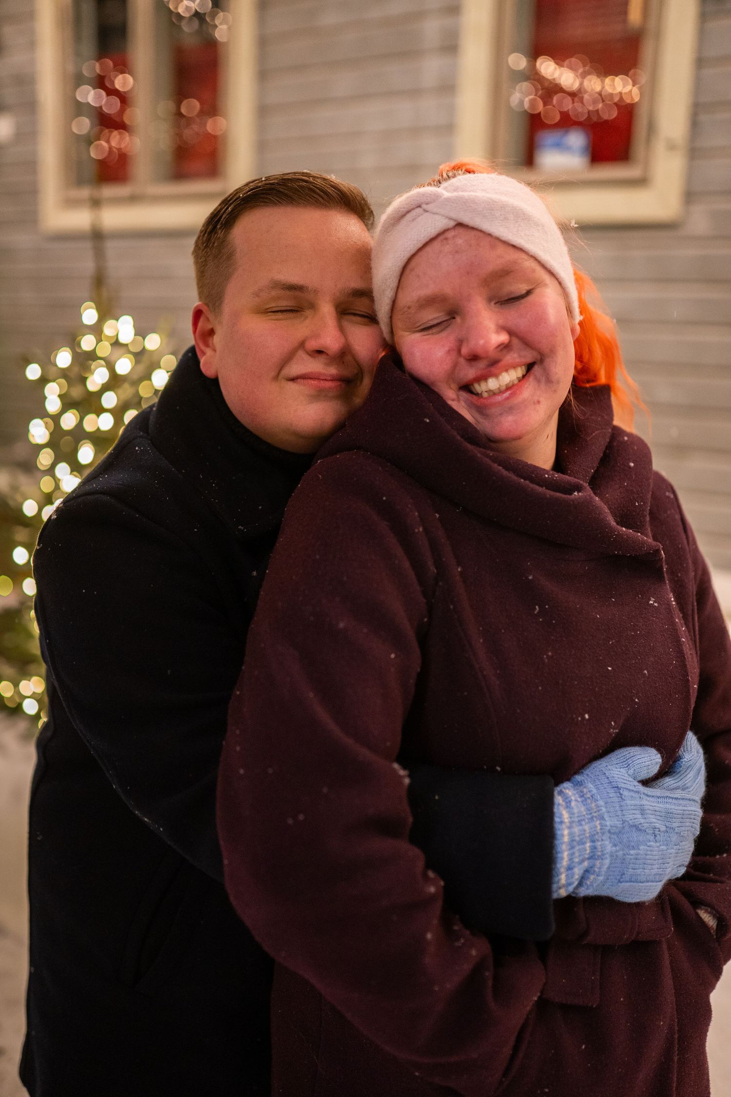 Two people embracing outdoors in winter, smiling warmly, with festive Christmas lights glowing in the snowy background.