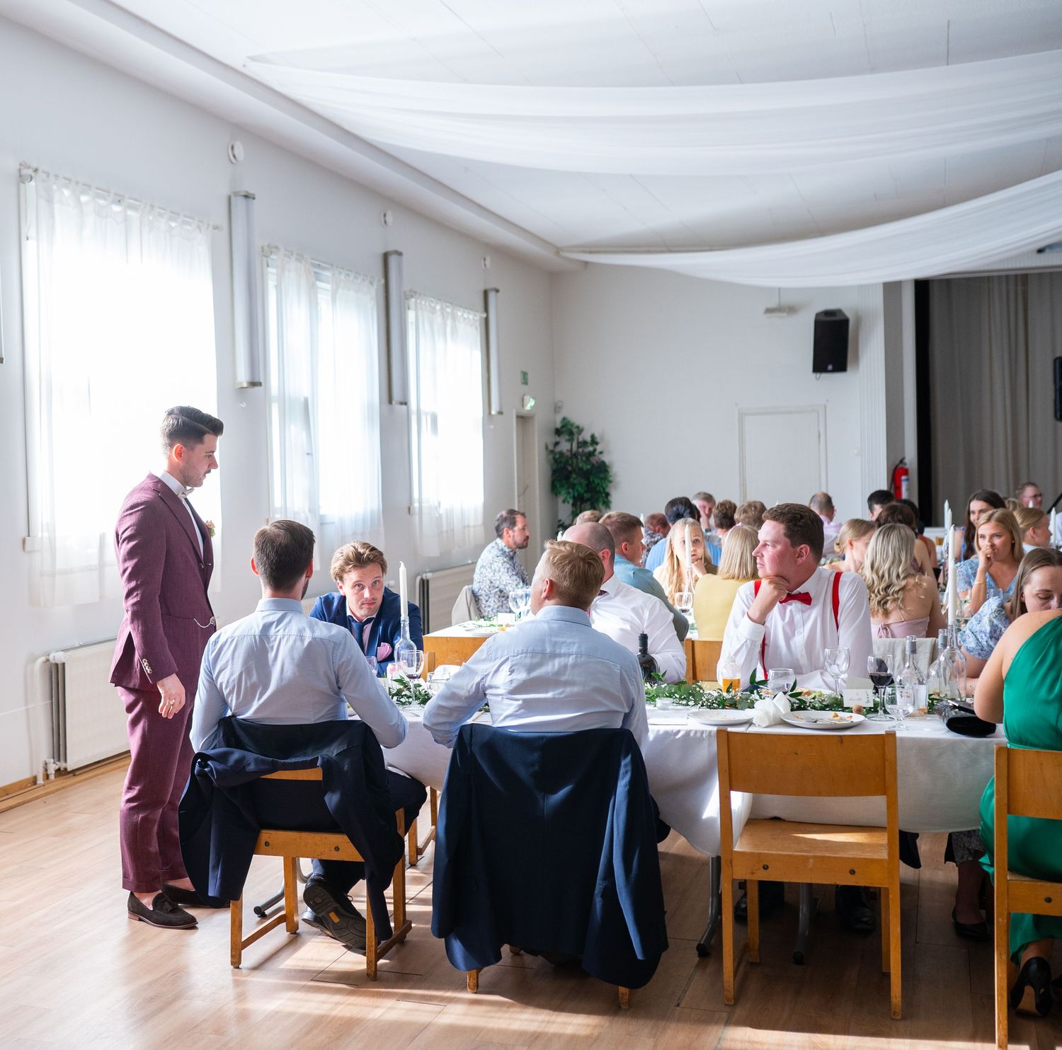 Wedding reception guests seated at long tables while groom in burgundy suit mingles in bright hall.