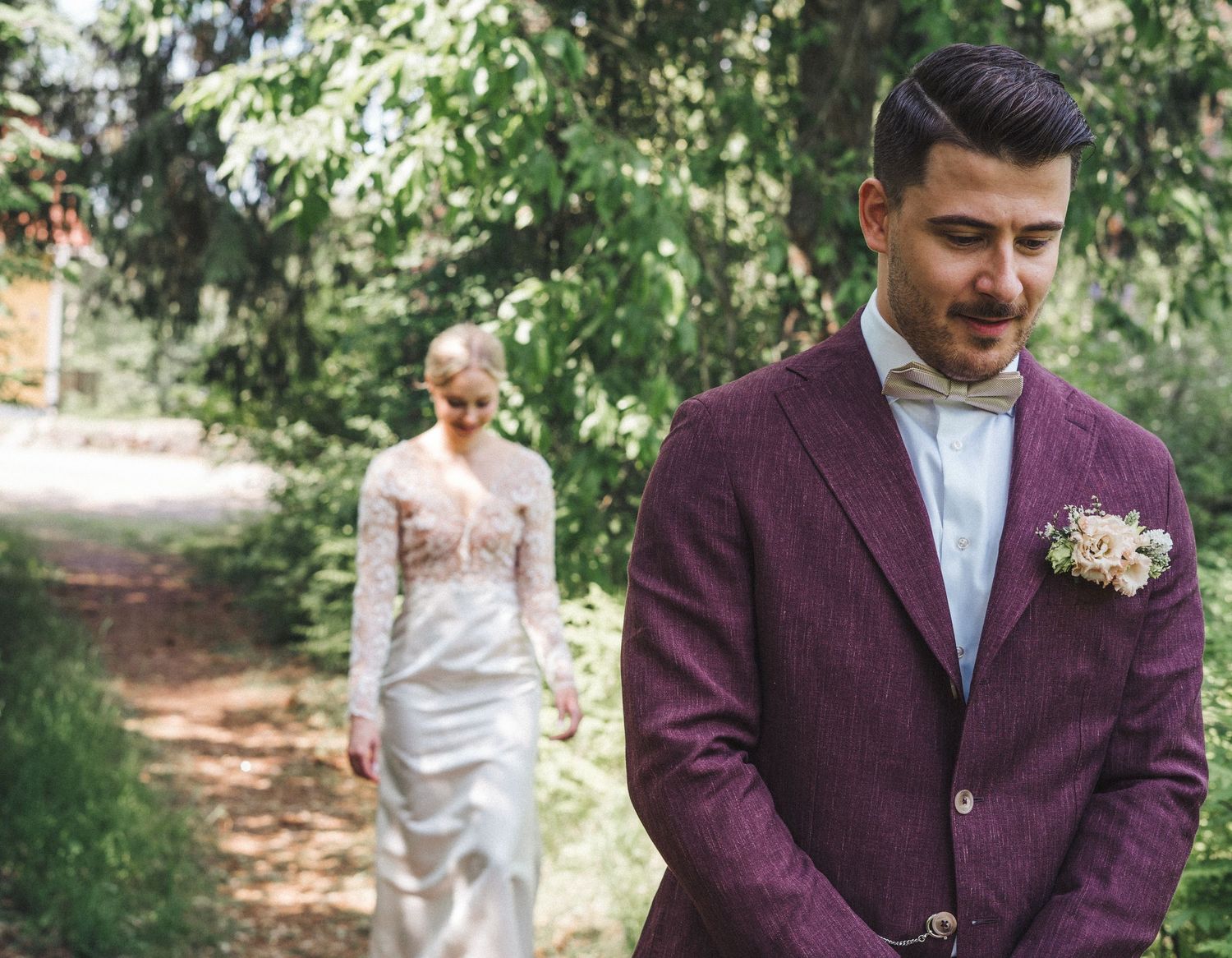 Groom in purple tweed suit with boutonniere stands as bride in lace gown walks toward him.