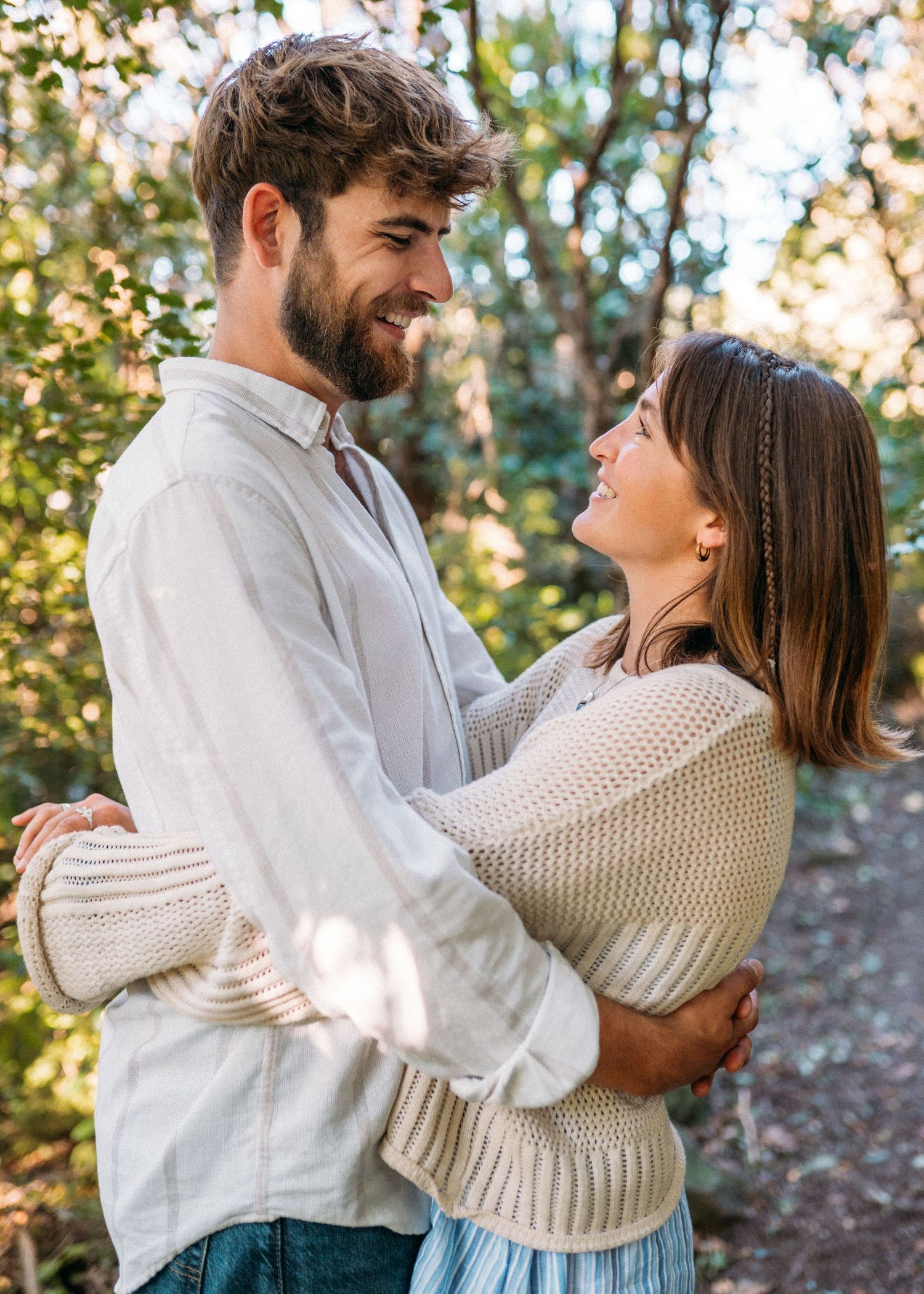 Happy couple gazing at each other while embracing on a sunlit forest path outdoors.