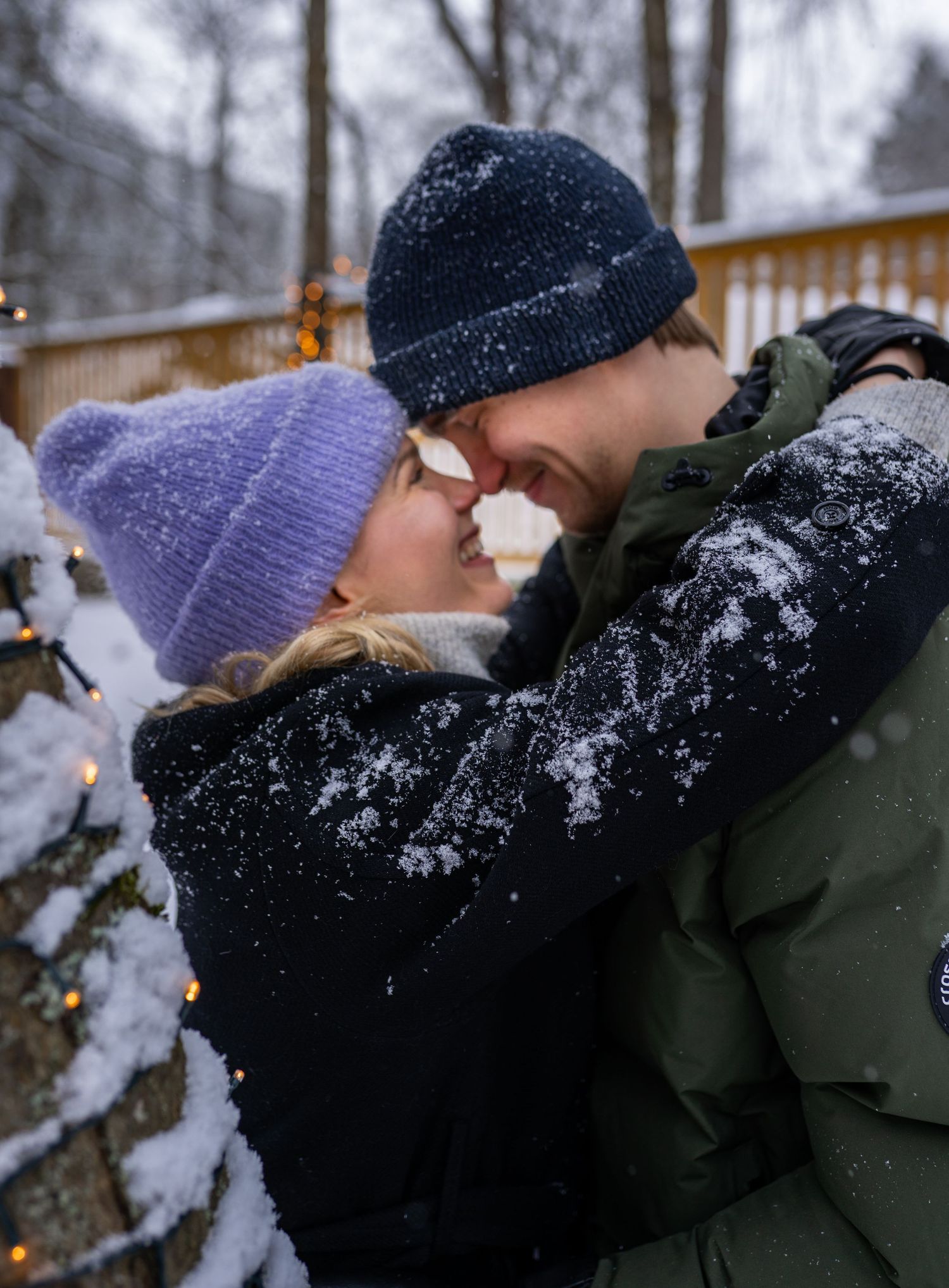 Couple sharing a romantic kiss outdoors in winter snow, wearing knit hats and coats near a snow-covered Christmas tree.