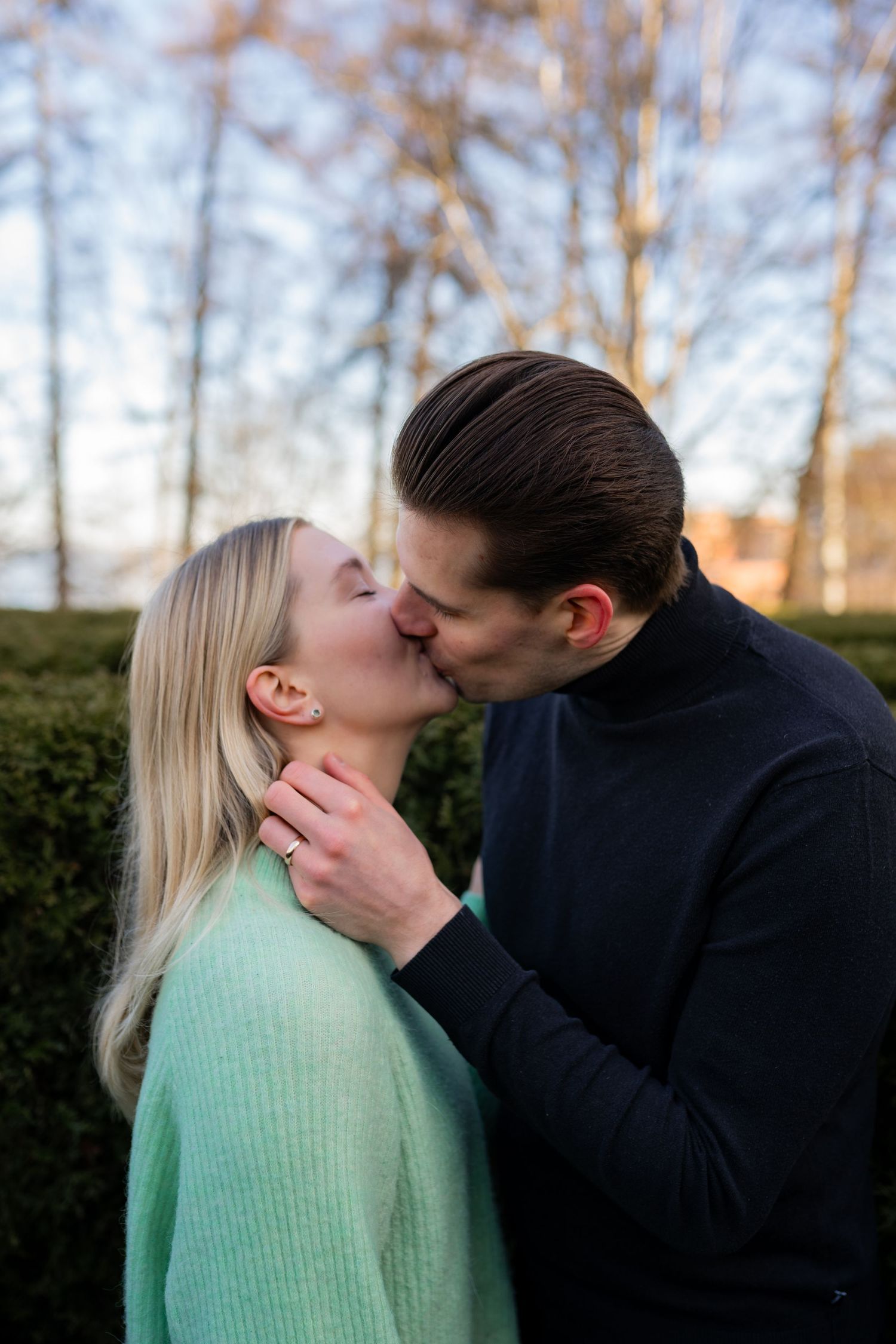 A couple shares a romantic kiss outdoors, with the woman in a mint sweater and man in a navy turtleneck, surrounded by greenery.