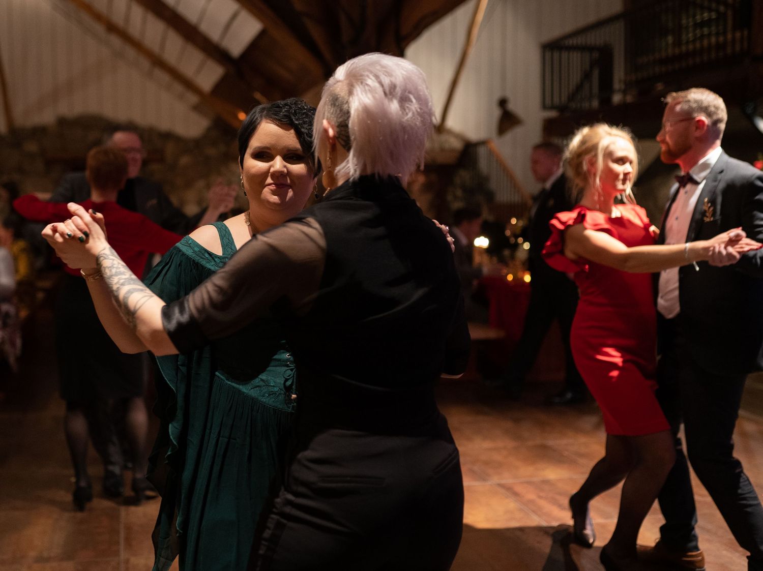 Adults dancing elegantly at a formal event, wearing green, black, and red dresses in a warmly lit venue.