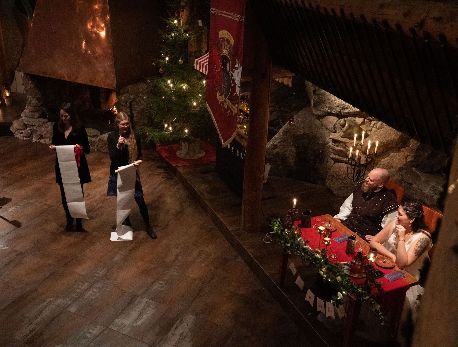 Medieval Christmas dinner scene with candlelit table, decorated tree, and guests in a rustic stone hall.
