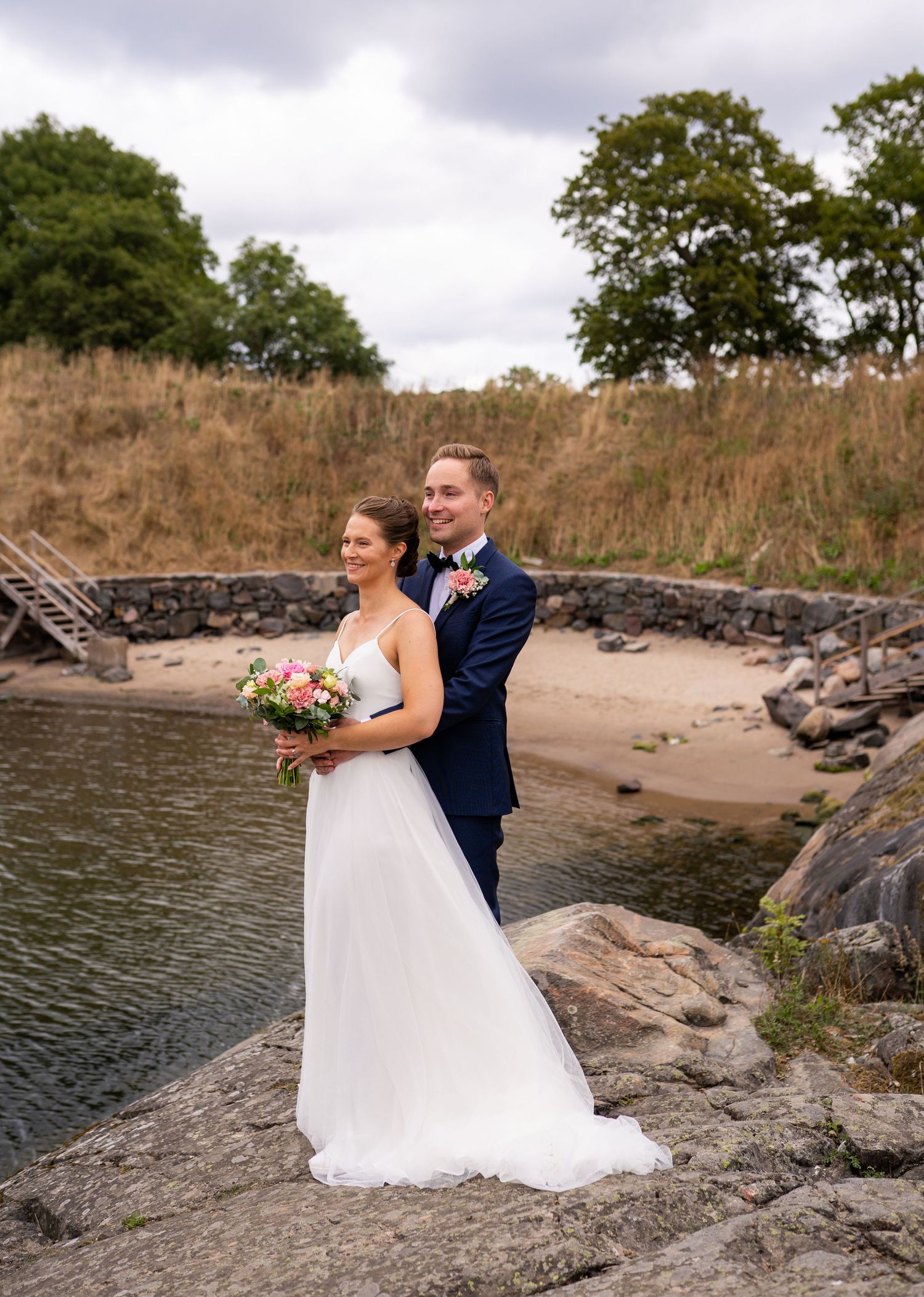 Bride and groom posing on rocky shore by calm water, she holds pink bouquet, he wears navy suit.