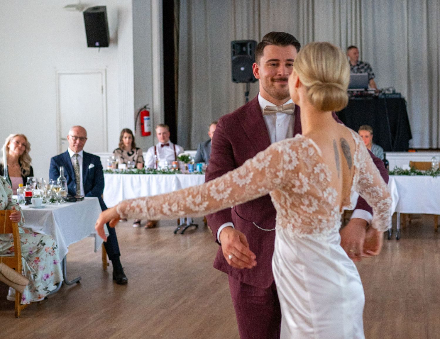 Smiling bride and groom share first dance at wedding reception while family watches from head table.