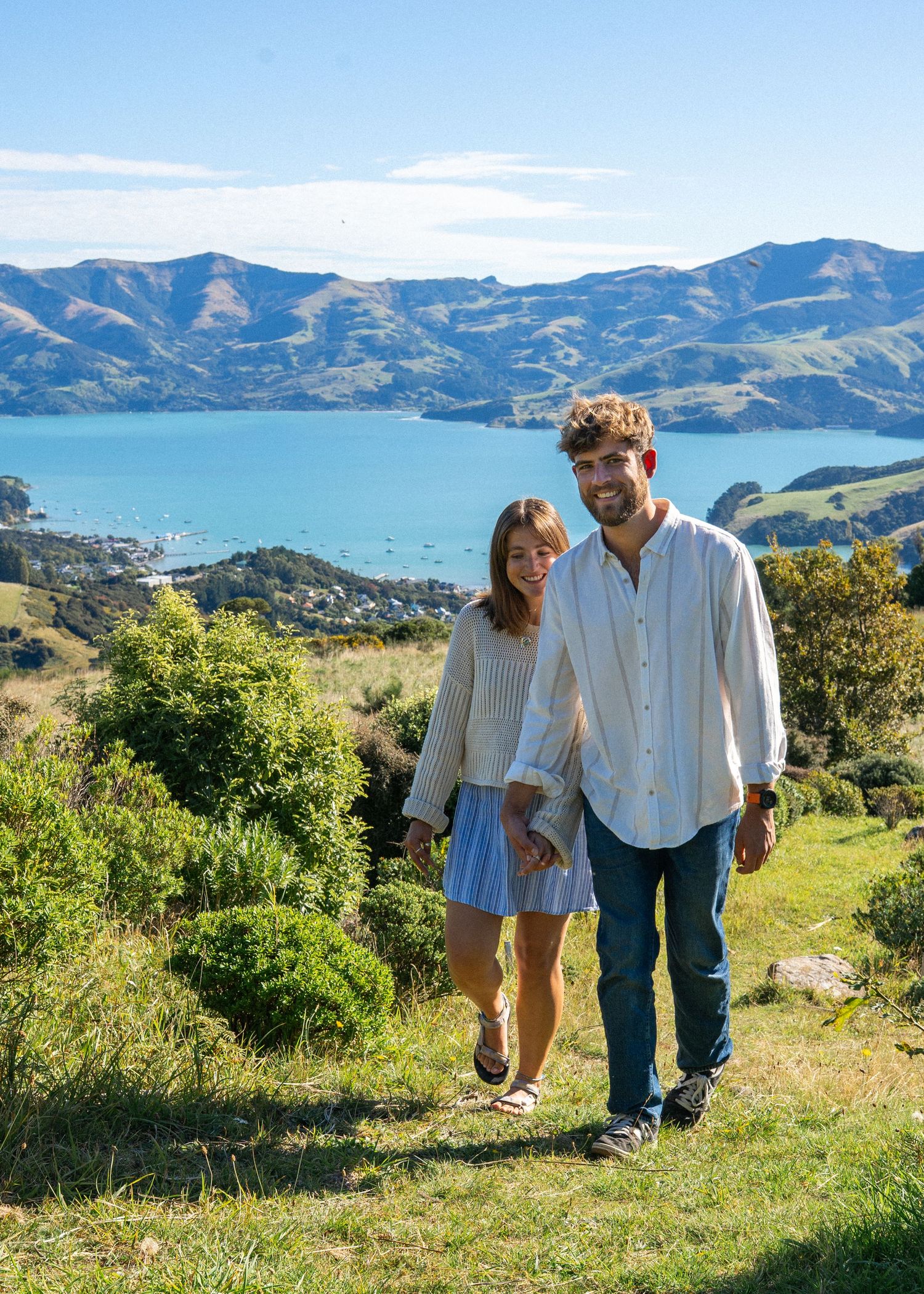 Happy couple walks hand in hand on grassy hillside with scenic blue harbor backdrop.