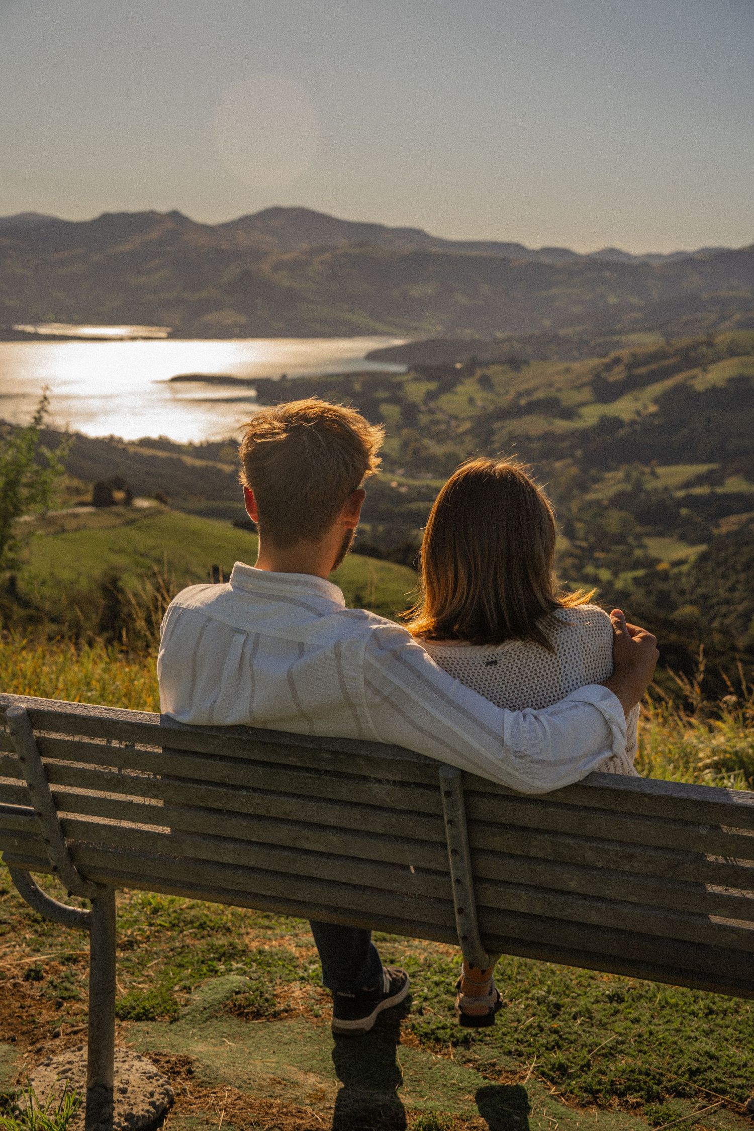 A romantic couple sits on a bench overlooking a stunning coastal valley and sparkling water at golden hour.