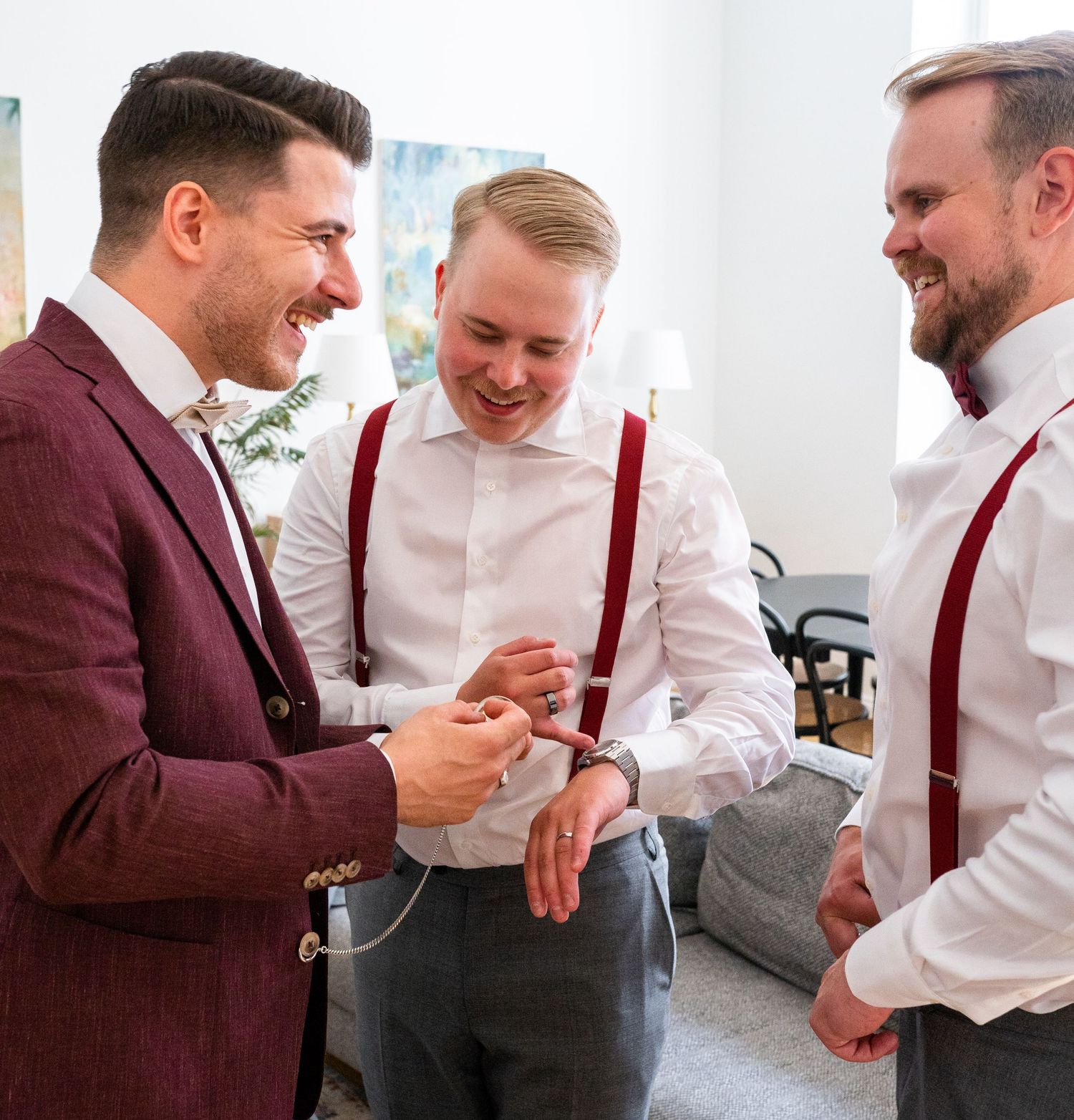 Groom and groomsmen in burgundy suspenders laughing together while getting ready for a wedding.