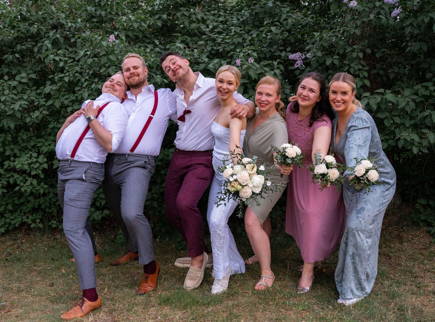 Wedding party posing playfully outdoors with bouquets, wearing burgundy and pink attire against green foliage.