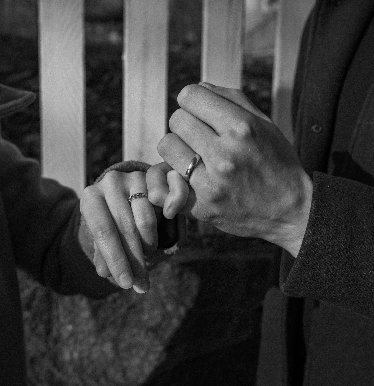 Black and white photo of two people holding hands, both wearing rings, standing near a wooden fence.