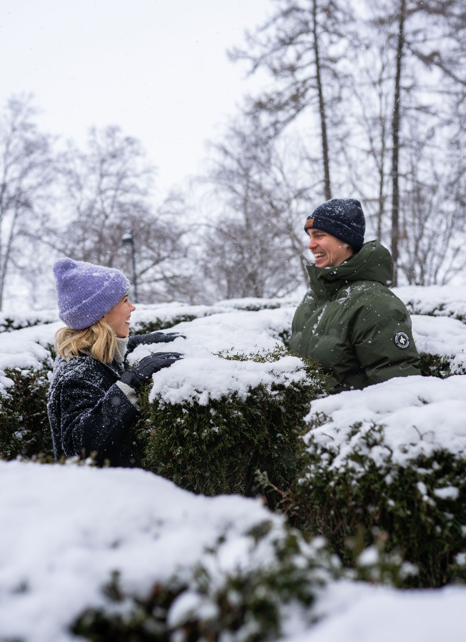 Two people in winter outerwear chat over snow-covered hedges in a snowy park setting.