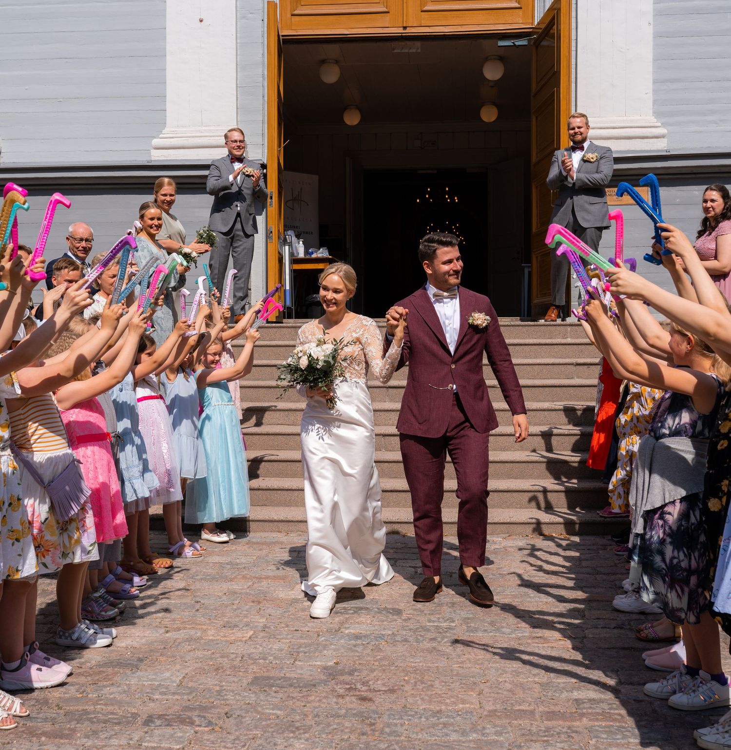 Bride and groom exit church steps as guests celebrate with colorful ribbon wands at their wedding.