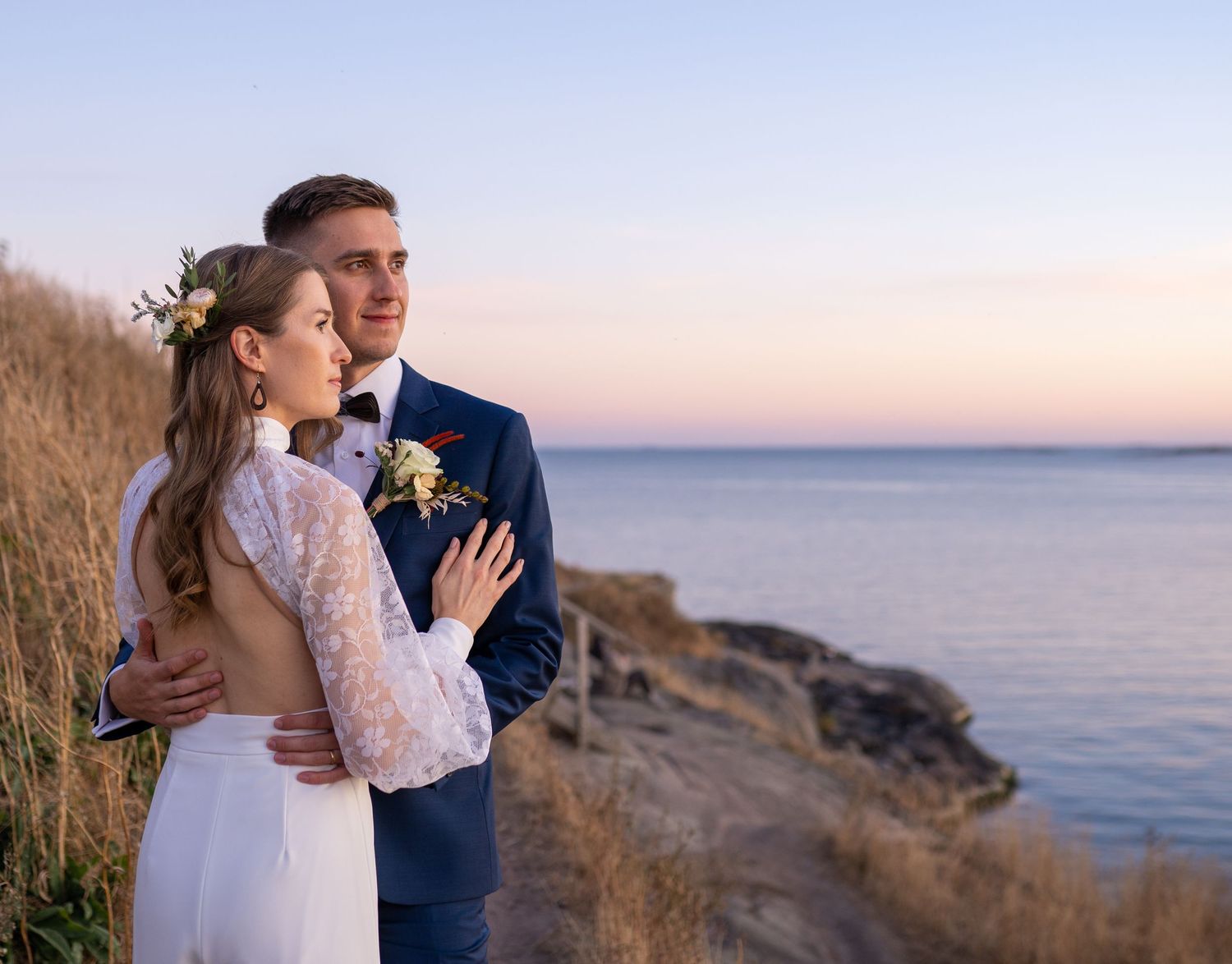 Romantic couple embraces at golden hour on coastal cliffside, bride wearing floral crown.