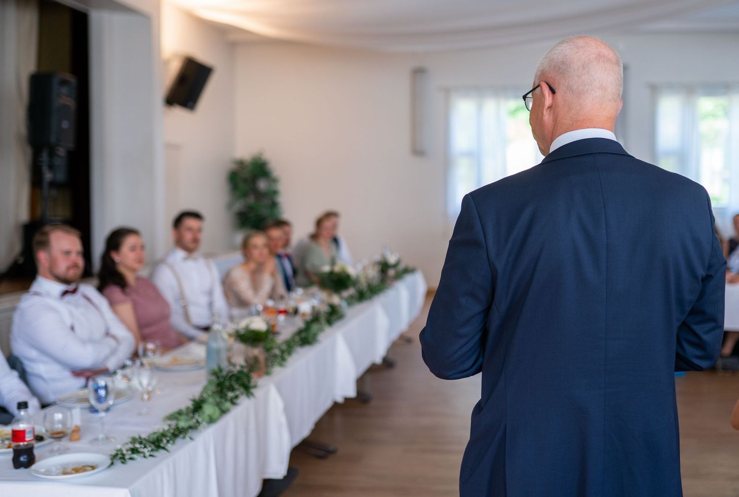 Bald man in navy suit giving a heartfelt speech to seated wedding guests at a reception.