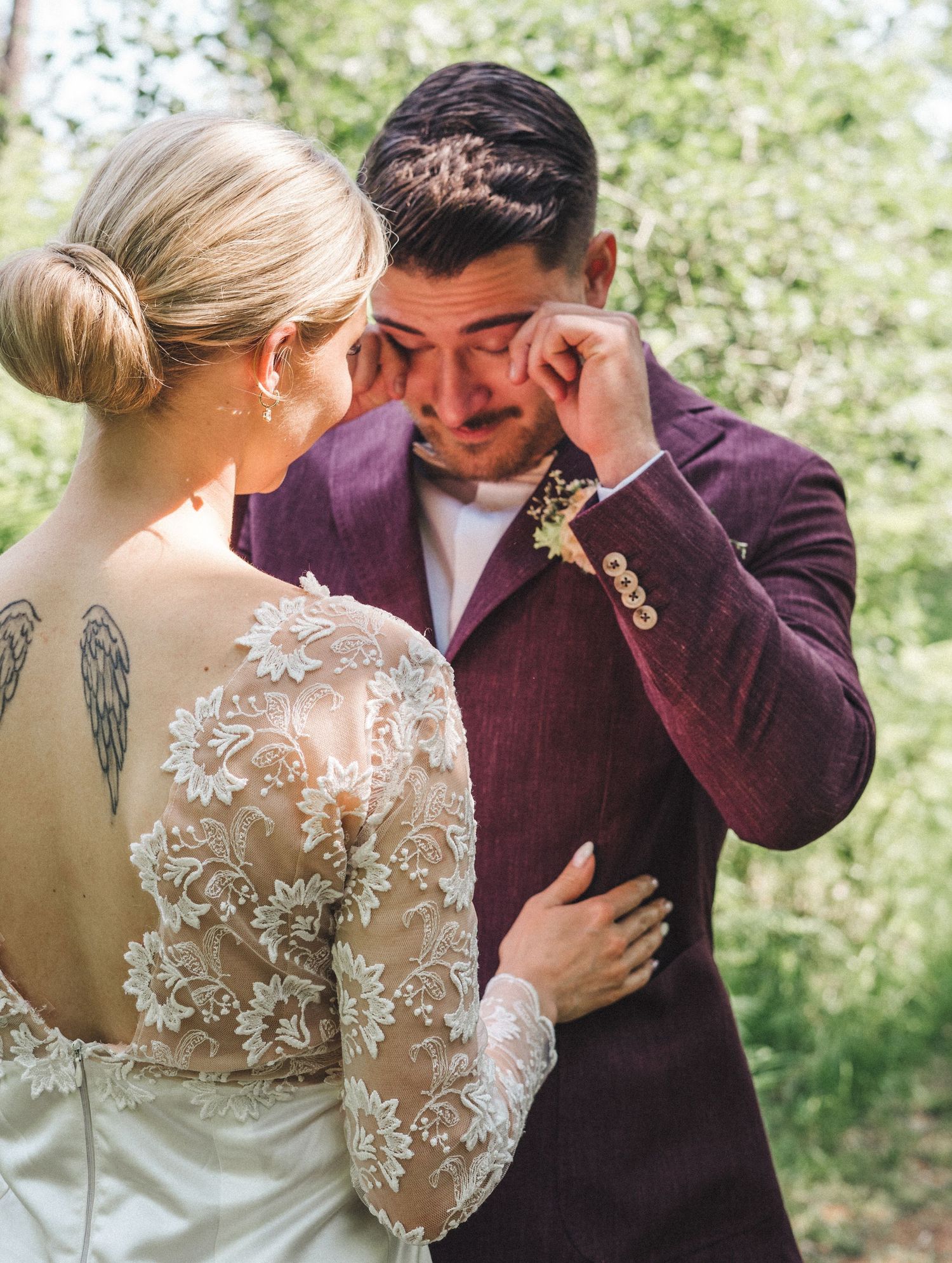 Tearful groom in purple suit wipes eye while embracing bride with angel wing tattoo in lace wedding gown.