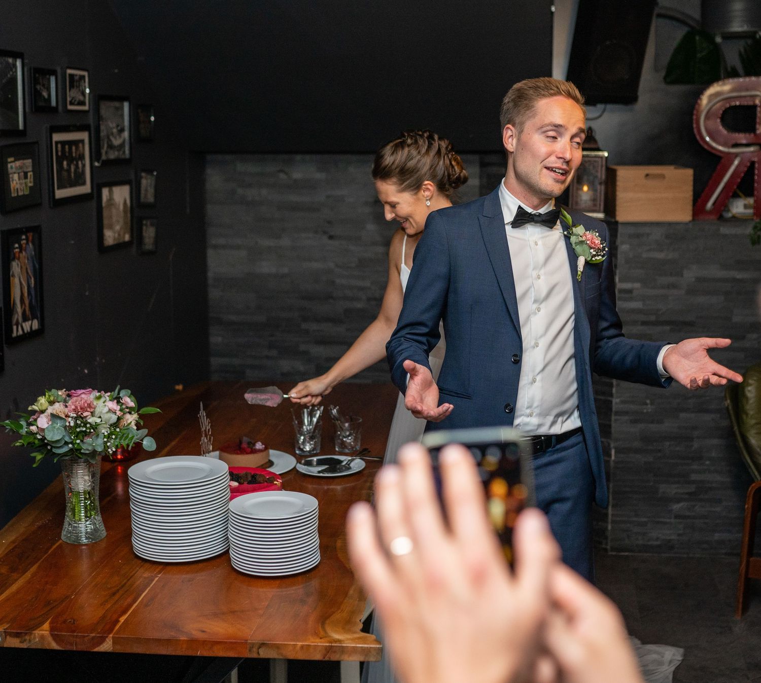 Groom gesturing expressively beside bride at wooden table during wedding cake cutting ceremony.