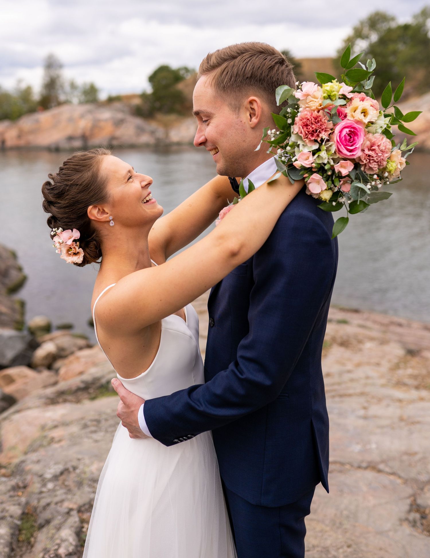 Bride and groom smiling at each other by rocky waterside, bride holding pink bouquet.