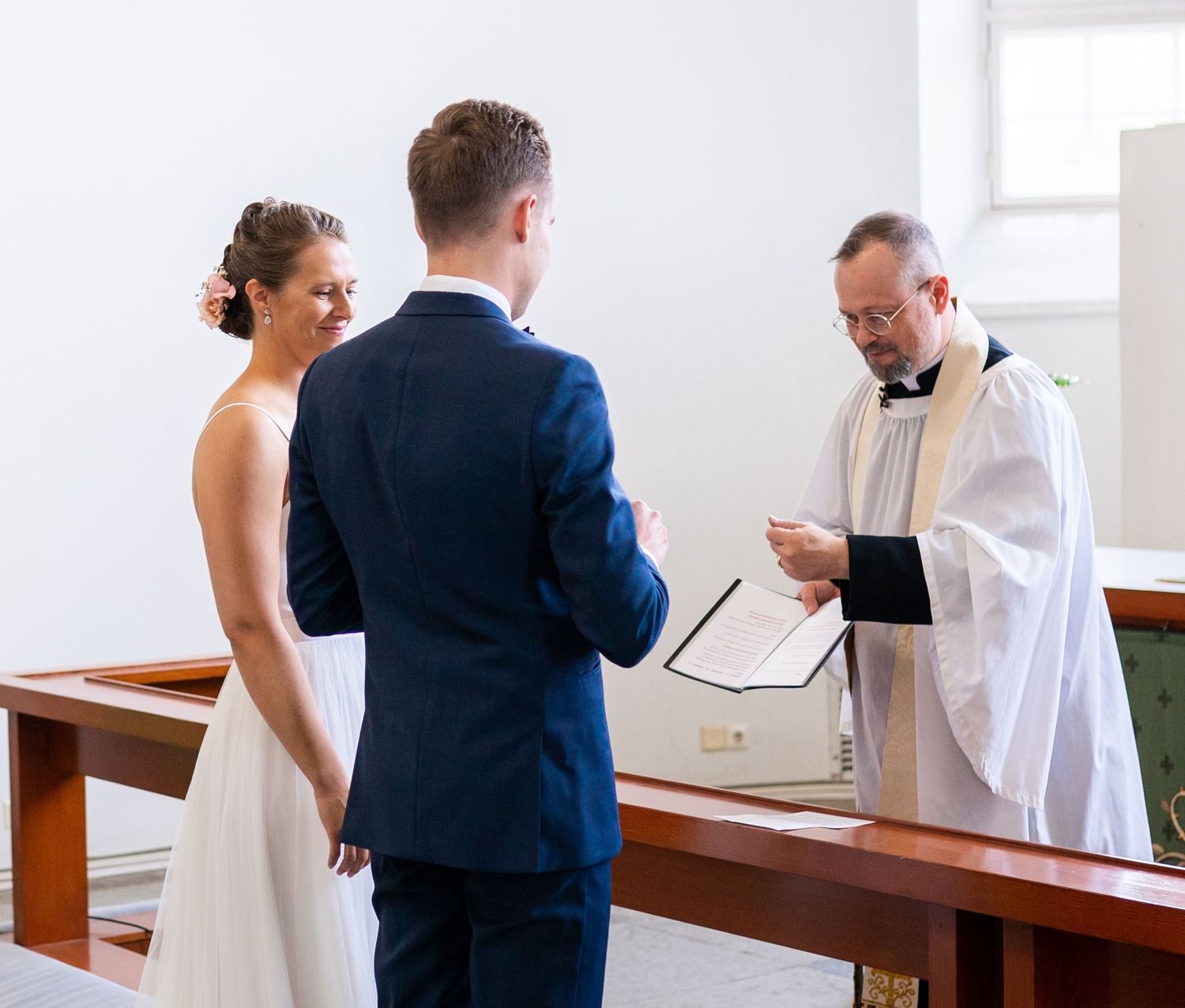 Smiling bride and groom exchanging vows with officiant at church wedding.