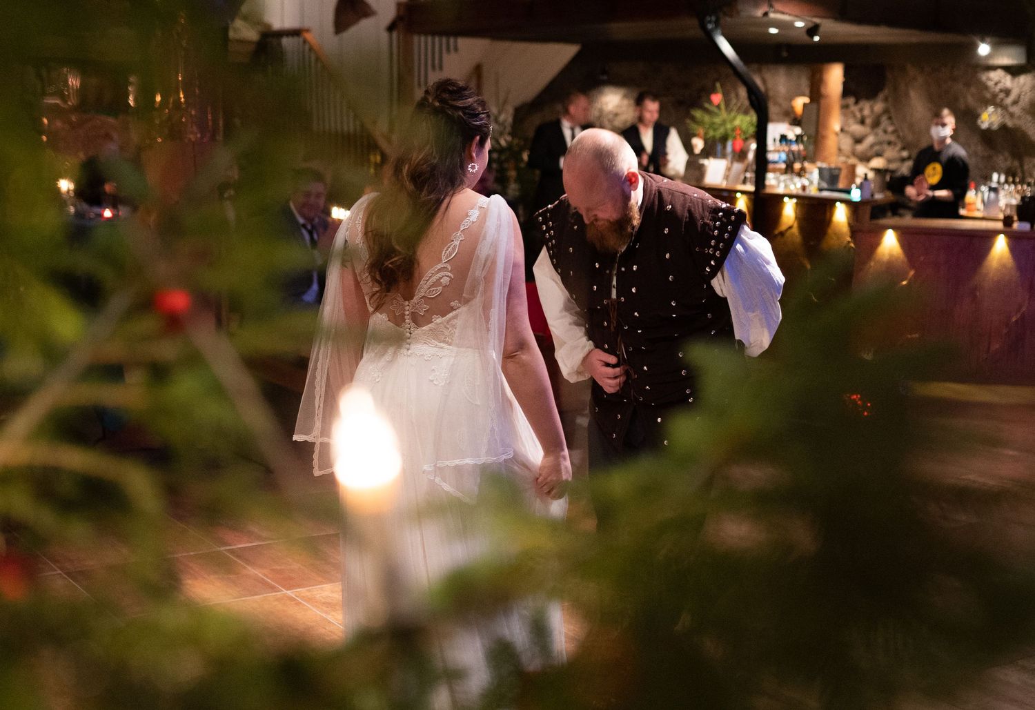 Couple in elegant attire dancing at a festive indoor wedding reception surrounded by Christmas trees.