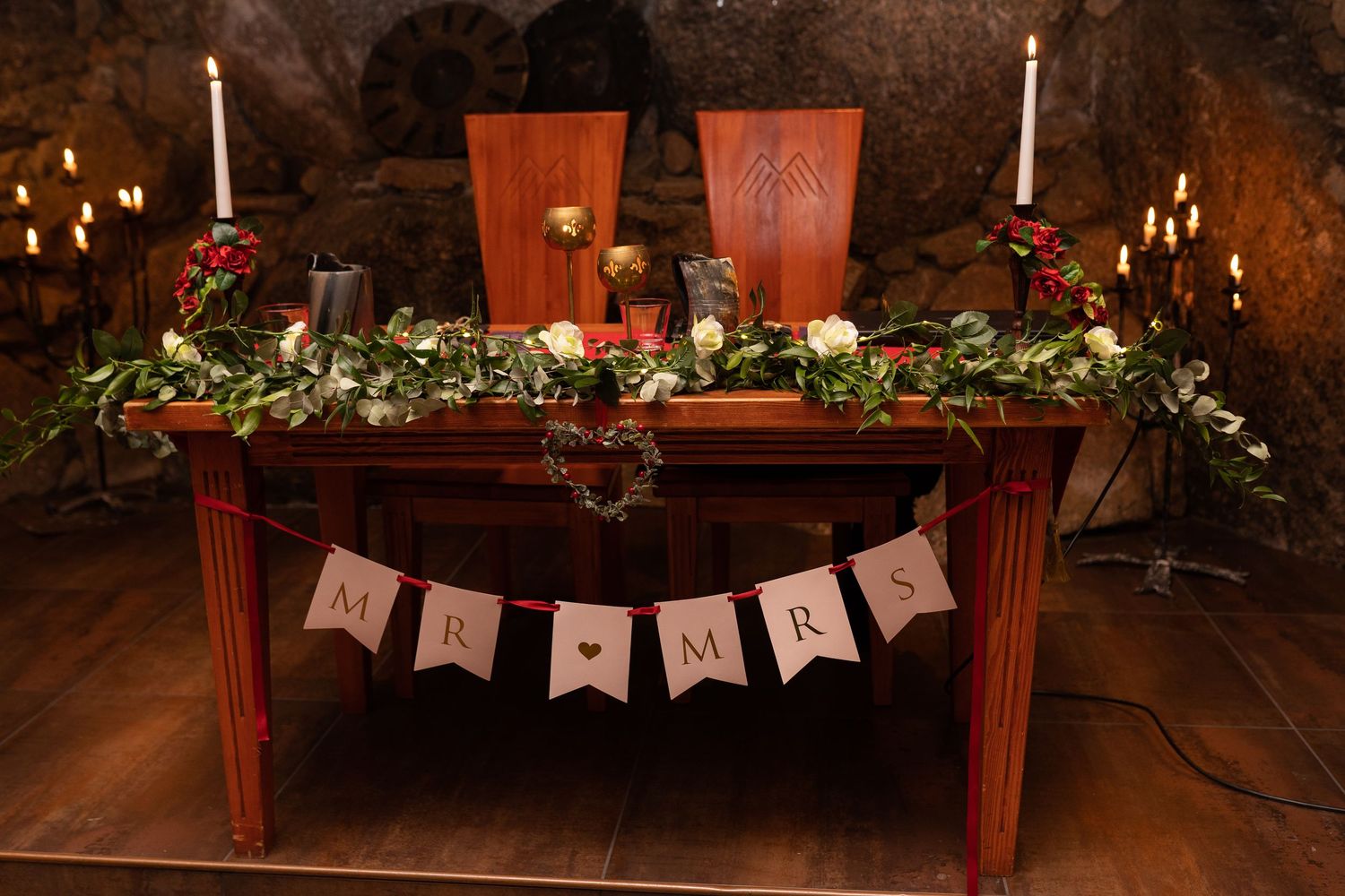 Rustic wedding sweetheart table with MR & MRS banner, greenery garland, white roses, and candles in a cave venue.