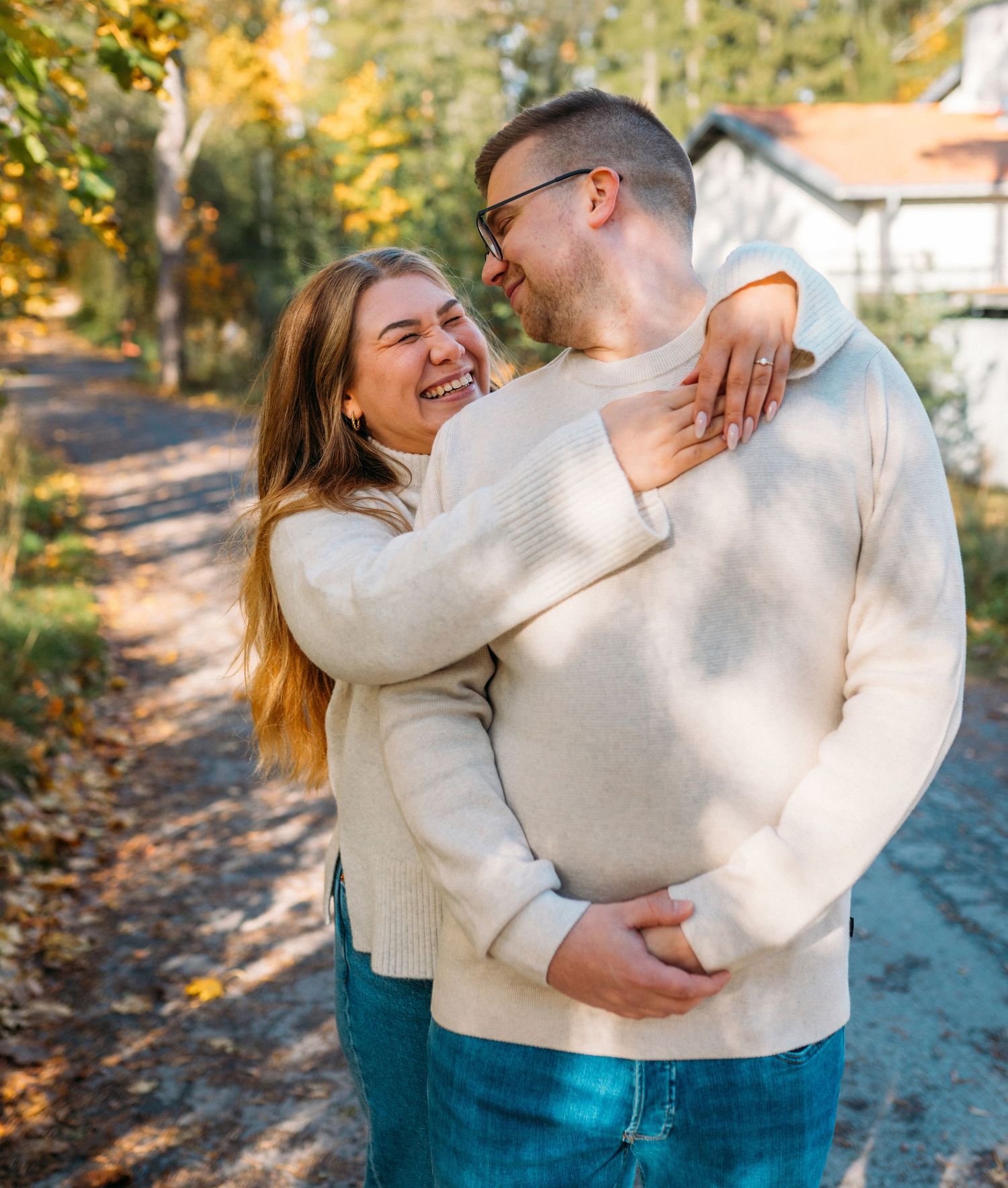 A smiling couple in matching cream sweaters and jeans embrace outdoors in autumn, surrounded by colorful fall foliage.