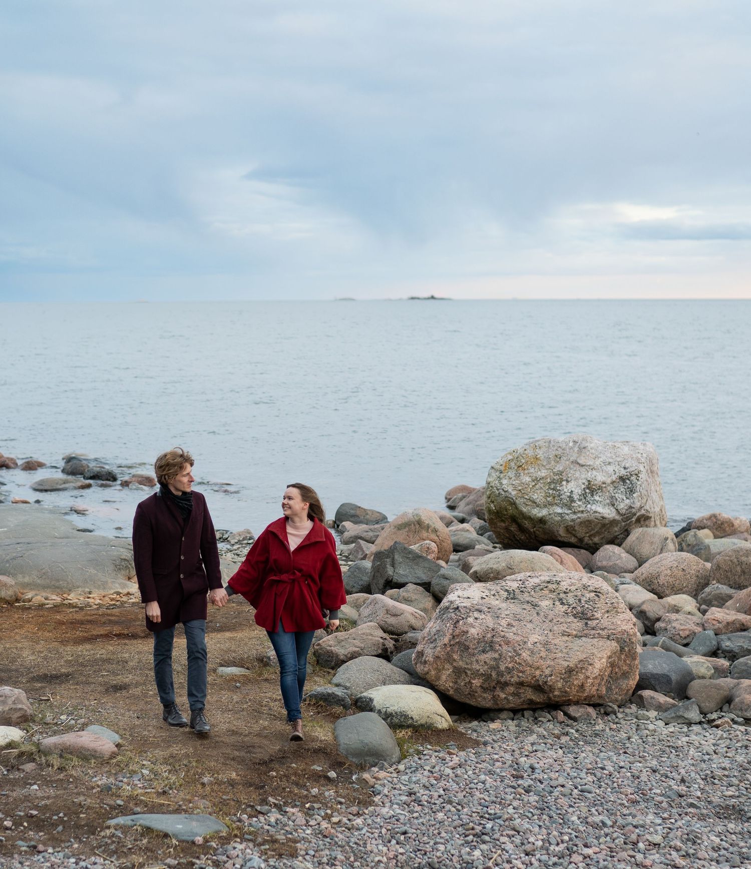 Couple holding hands walking along rocky shoreline with calm sea and cloudy sky.
