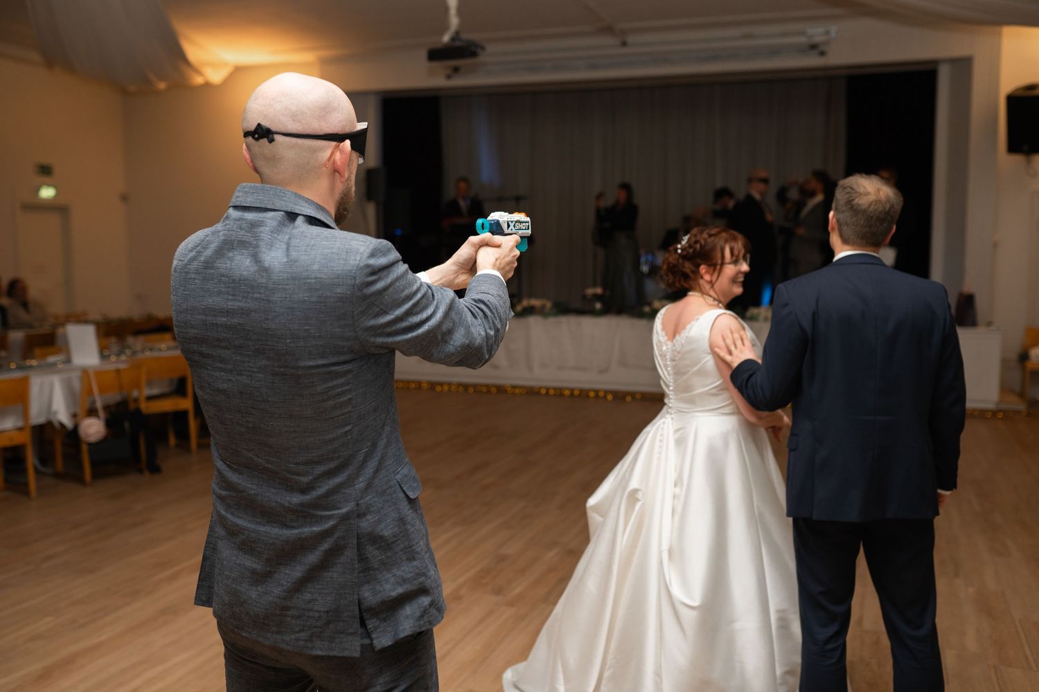 Wedding guest photographing bride and groom sharing first dance at indoor wedding reception celebration.