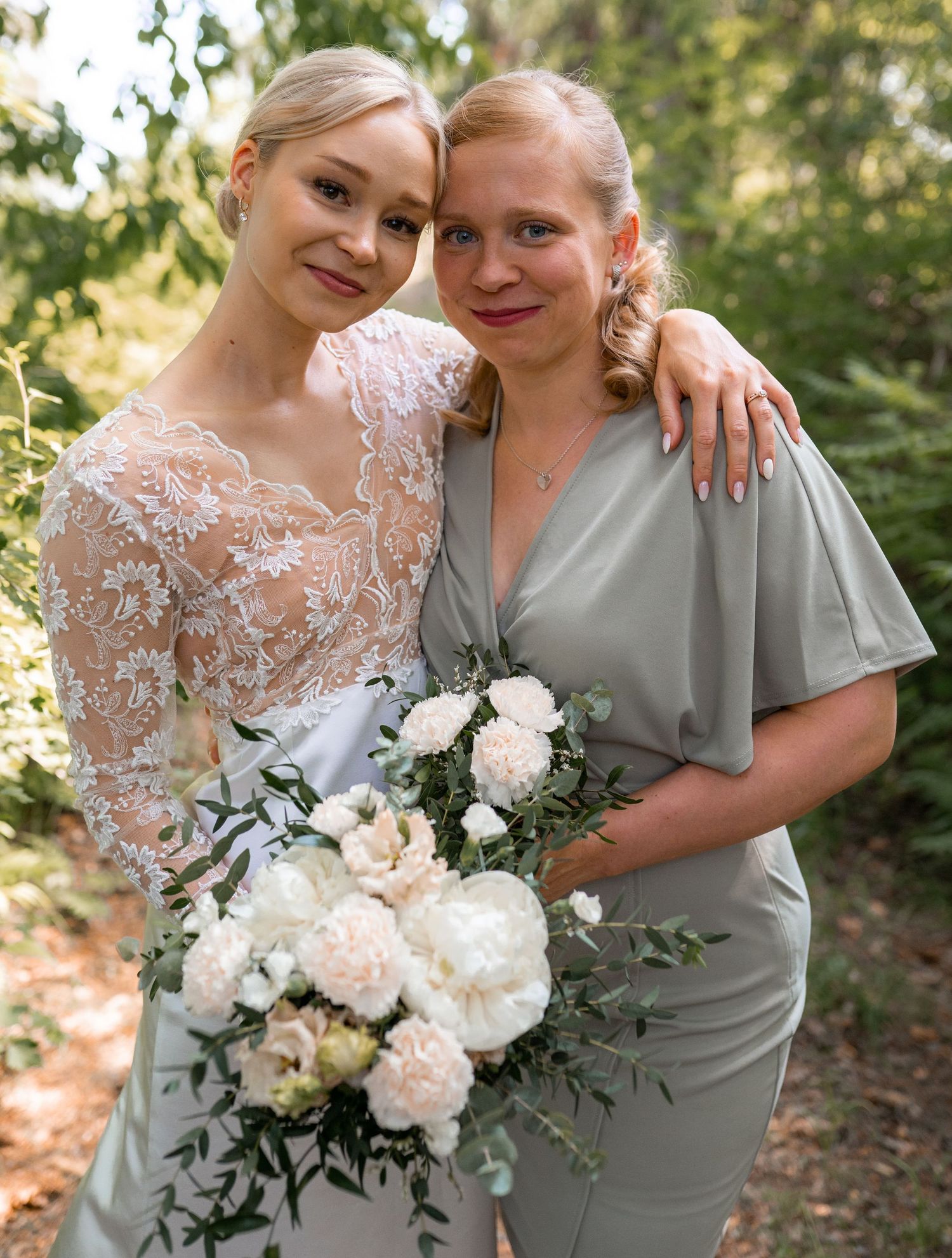 Bride in lace wedding dress posing with bridesmaid in sage green dress outdoors.