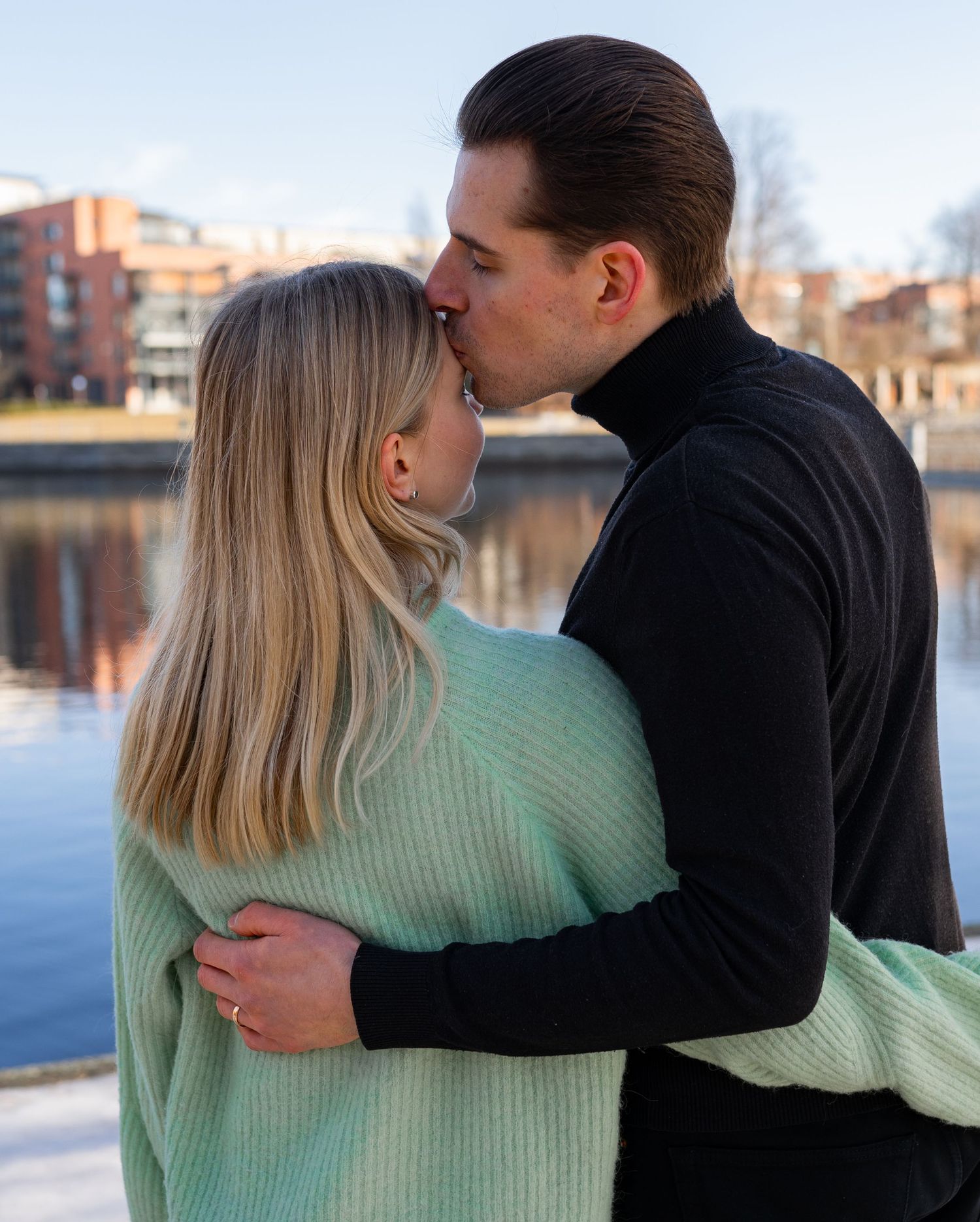 A couple sharing a romantic kiss outdoors by a river, woman in mint sweater and man in black turtleneck.