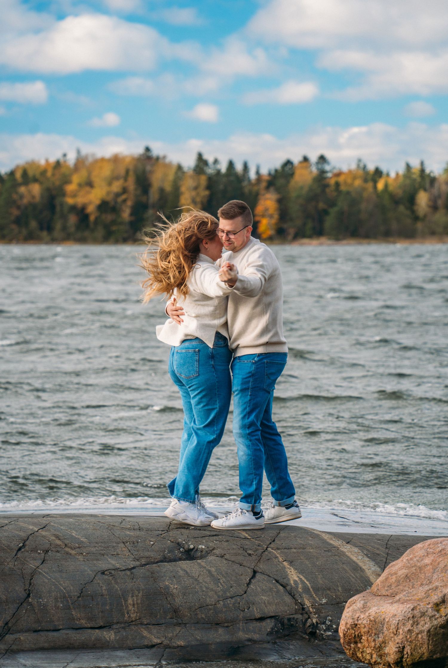 Couple dancing together on rocky shoreline with autumn trees and blue sky in background.
